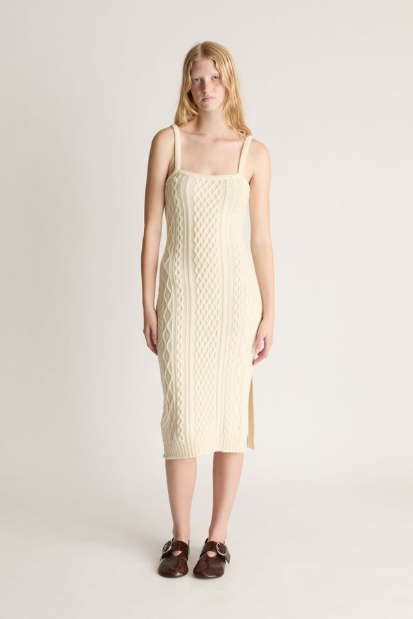 Woman wearing a cream cable knit midid dress paired with red shoes against a white studio background. 