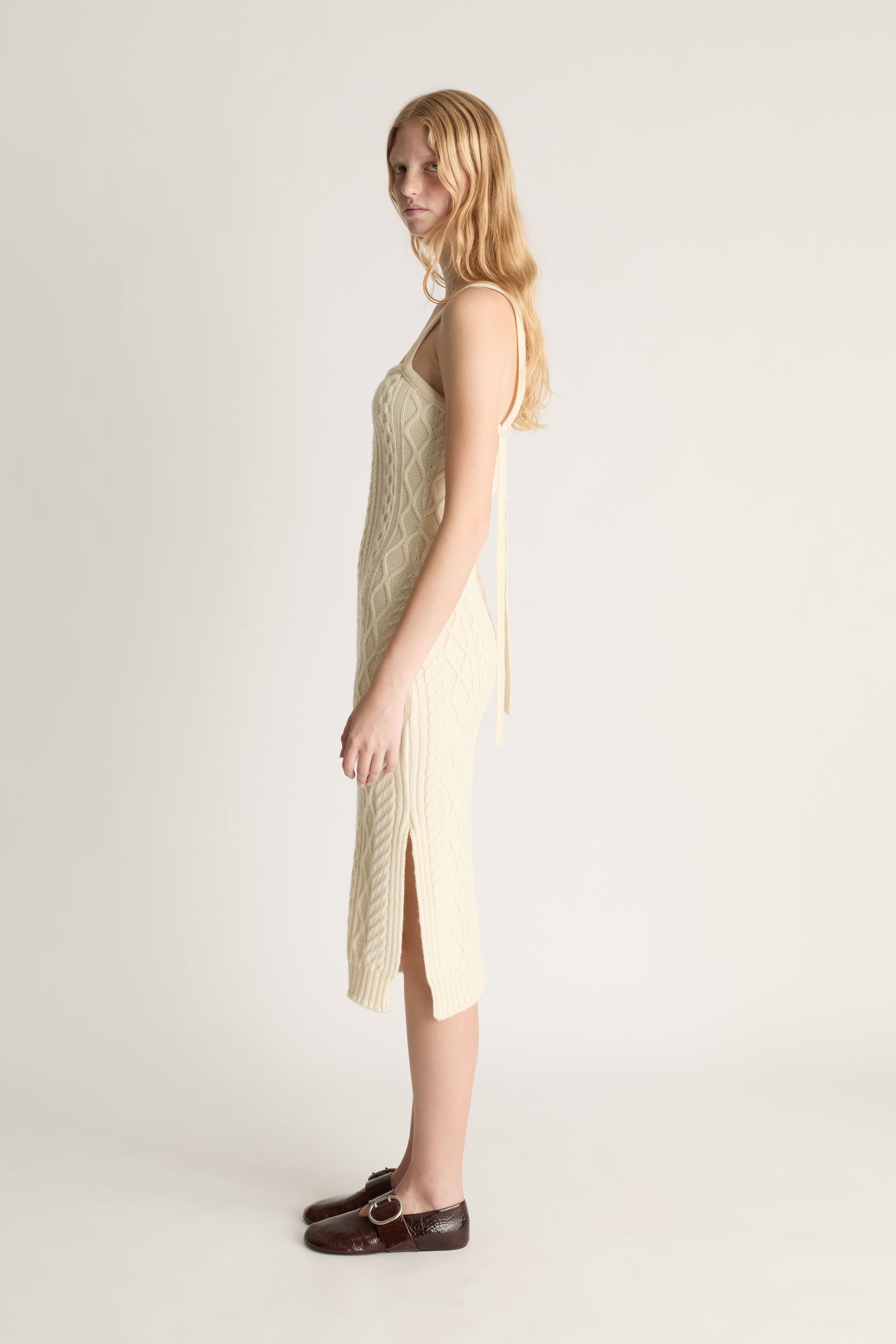 Profile view of a woman wearing a cream cable knit midi dress and red shoes shot against a white studio background. 