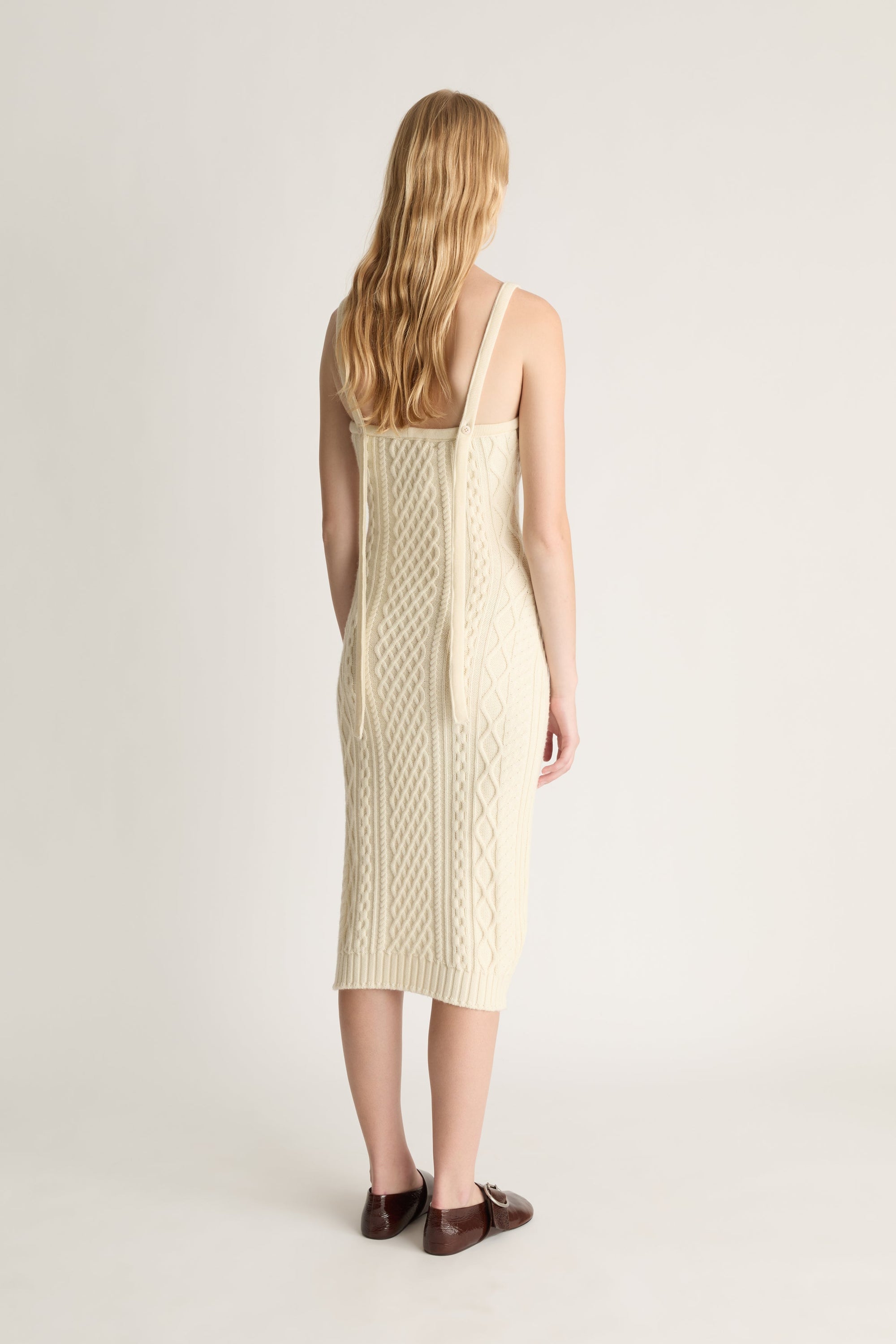 Back view of a woman wearing a cream-colored cable knit dress on on a white background