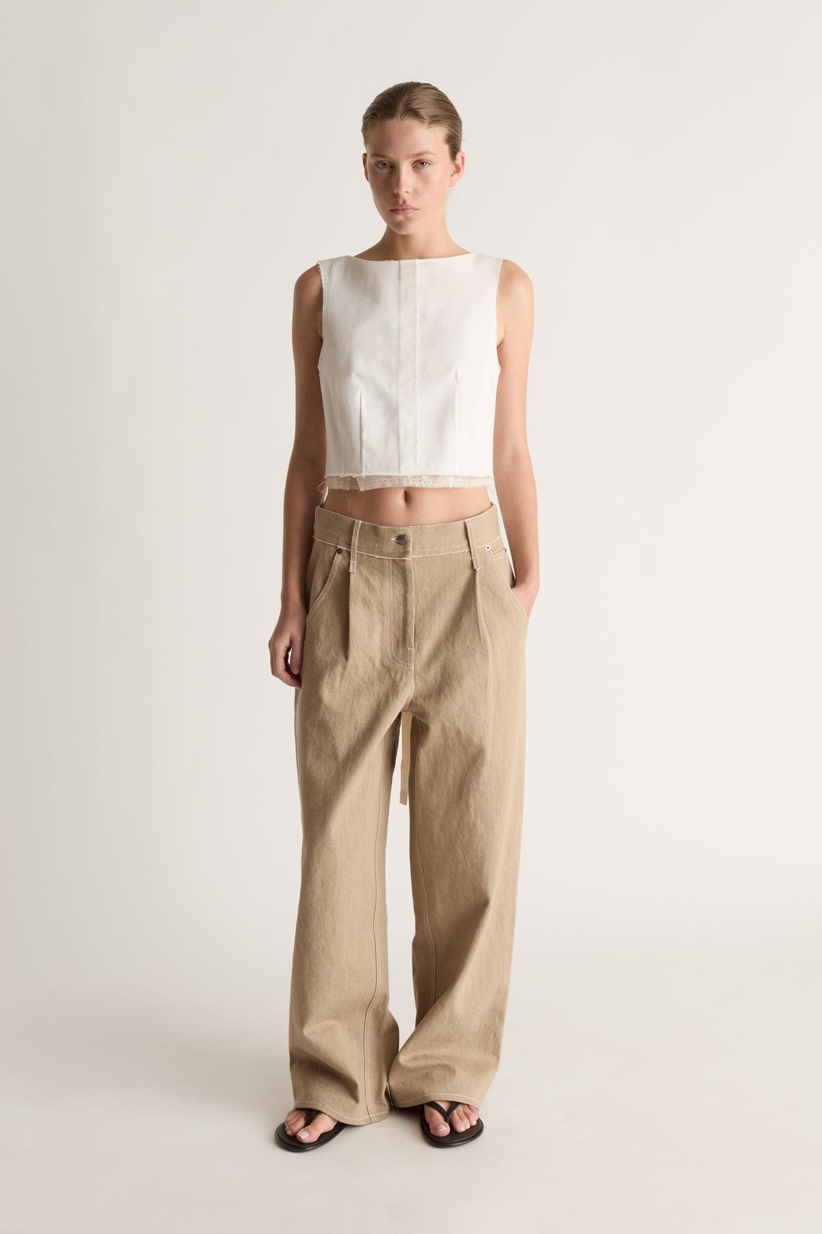 Front view of a woman with hair pulled back in a white panelled crop top, loose beige trousers and black flat sandals