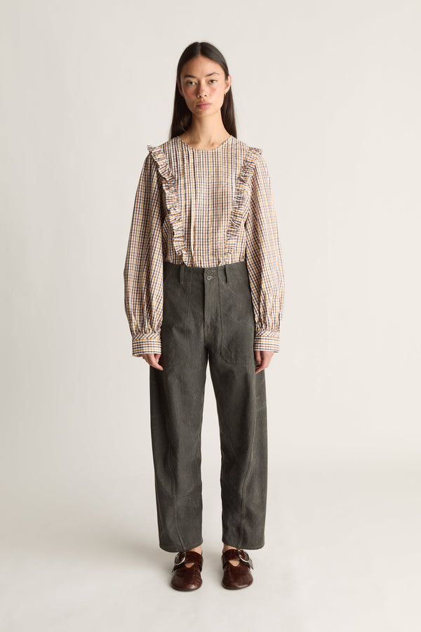 A model stands against a light gray studio background wearing a beige and brown checkered blouse with ruffles and long sleeves, paired with dark gray wide-leg trousers and brown shoes.