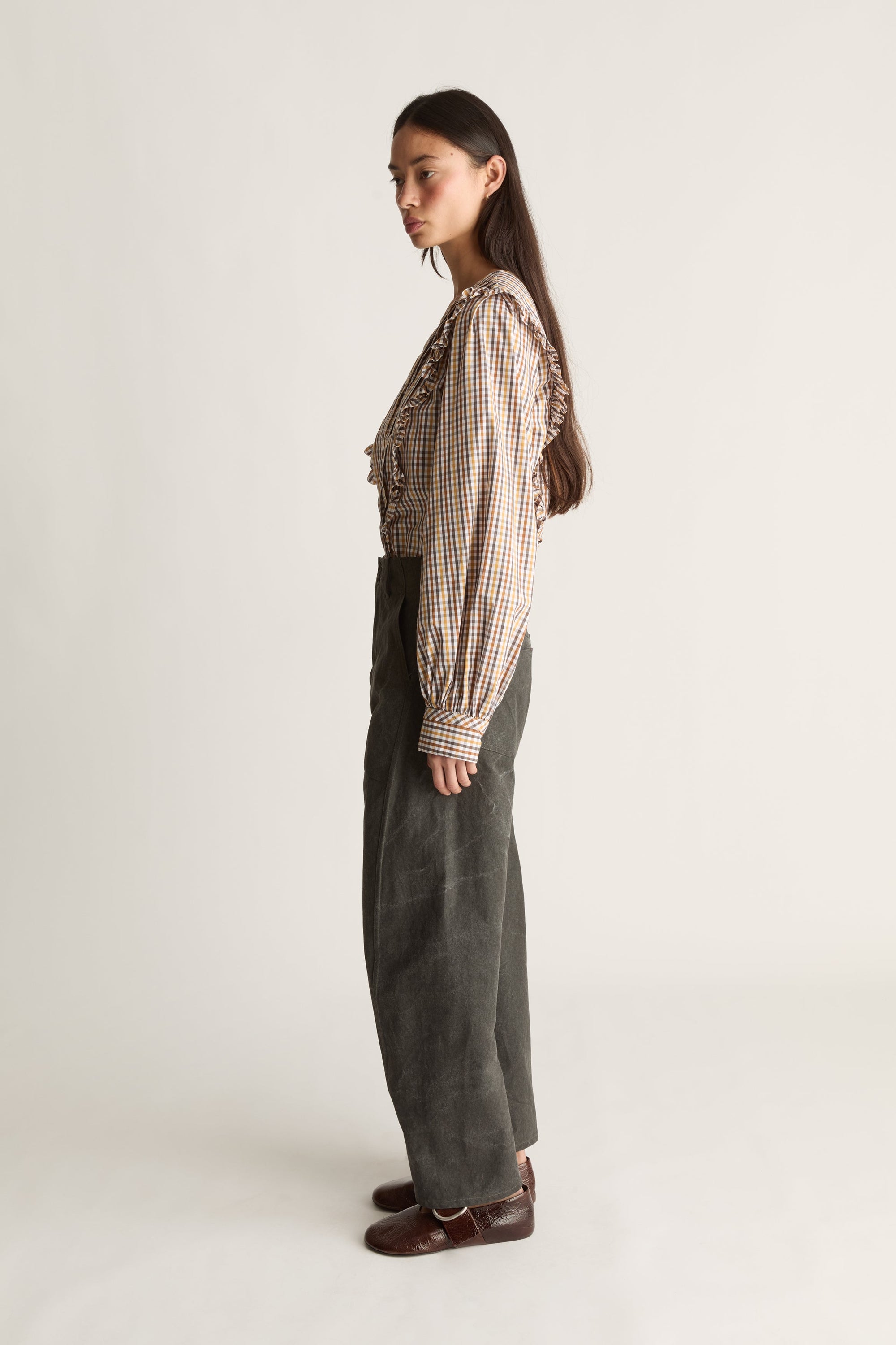 A woman stands in profile wearing a beige and brown gingham long-sleeve blouse with ruffled trim, paired with dark gray wide-leg trousers and brown leather shoes, against a plain light gray studio background.