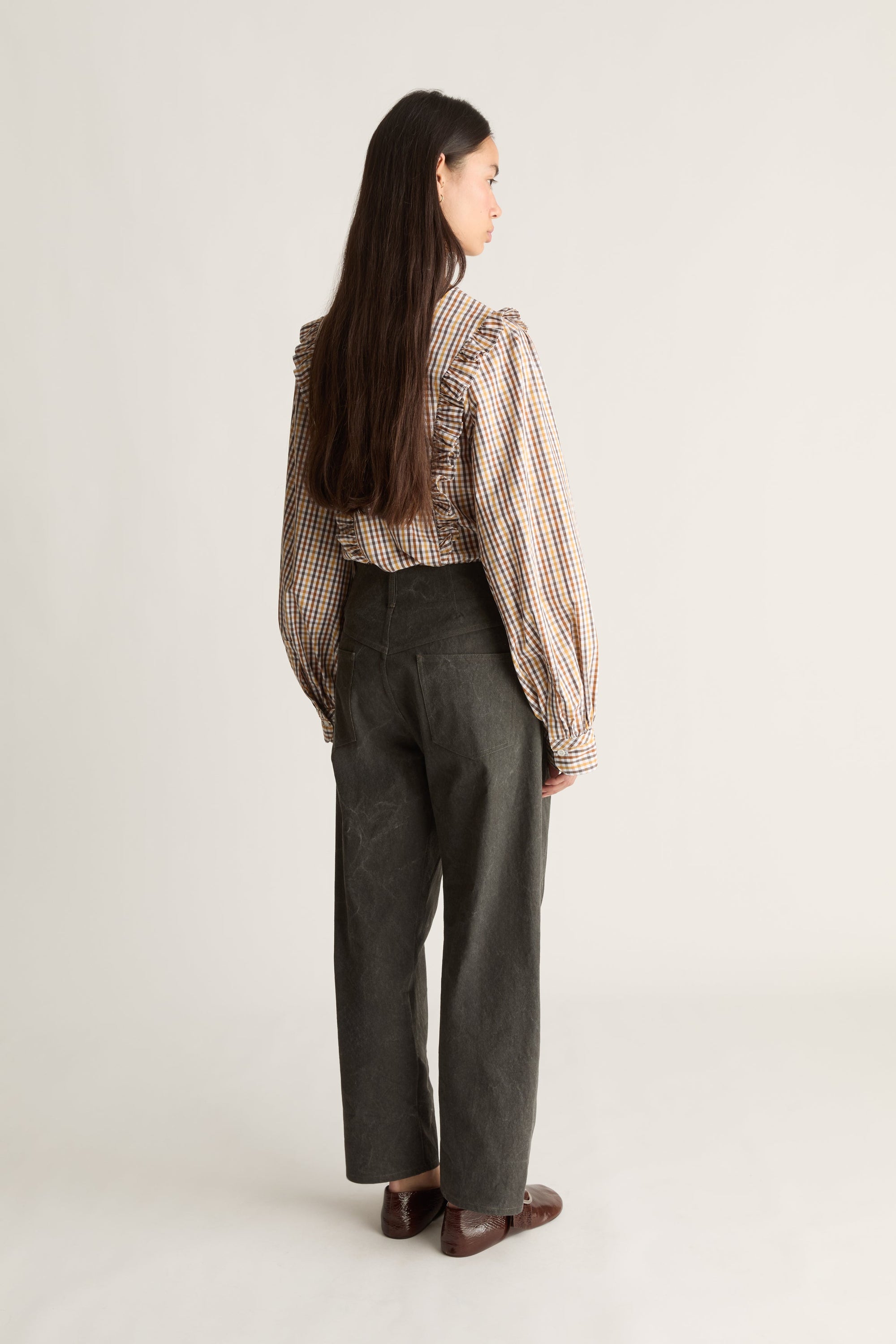 A woman is shown from the back wearing a brown and beige checkered long-sleeve blouse with ruffled shoulders and dark gray wide-leg trousers, standing against a plain light gray studio background.