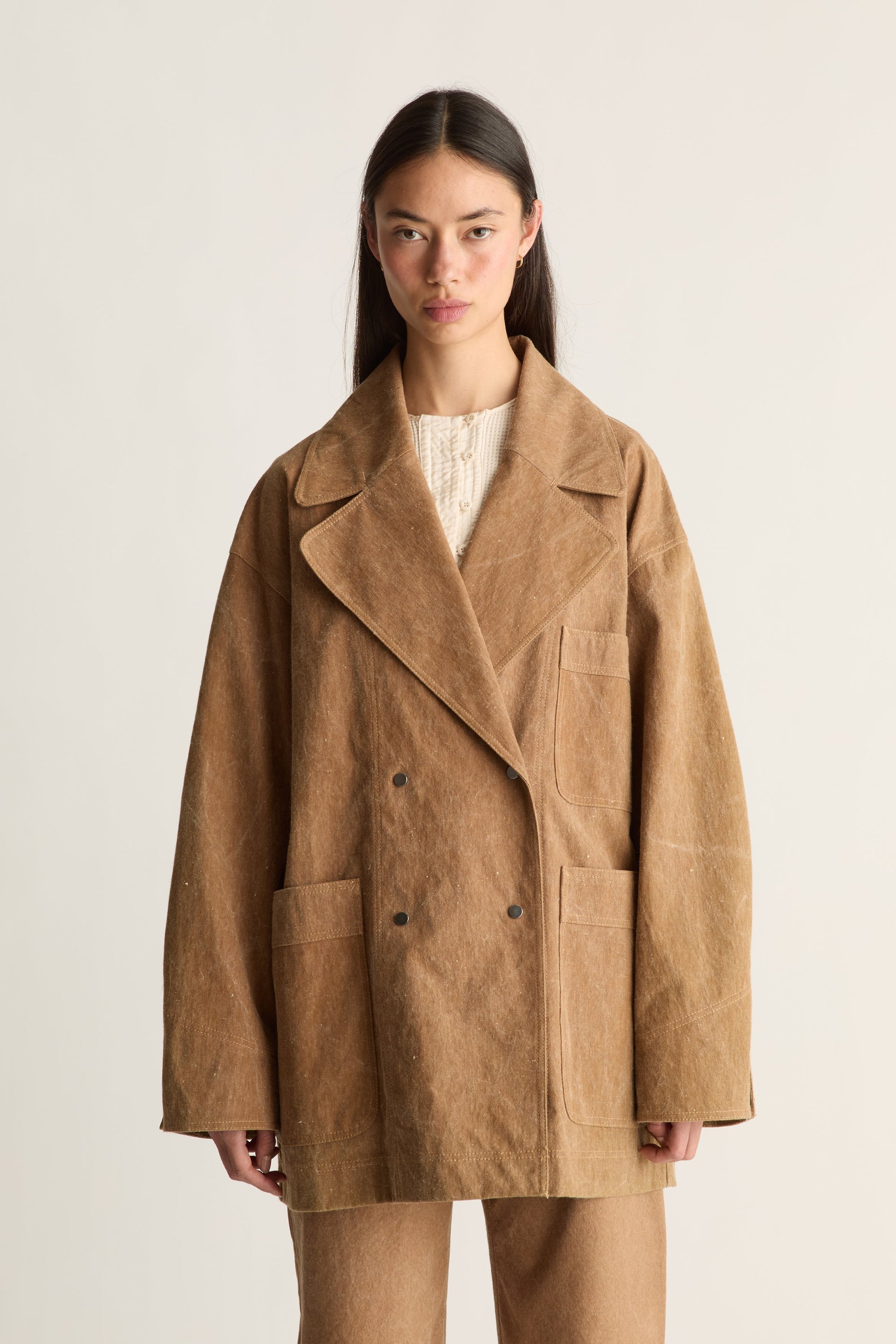 Close up of a woman wearing a brown coat and pants on a white background, facing the front.