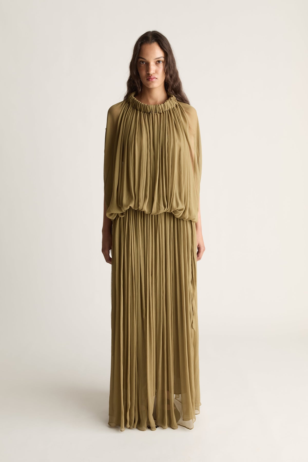 A woman wearing a floor length khaki dress that features a gathered neckline and a gathered waistband shot against a white studio background. 
