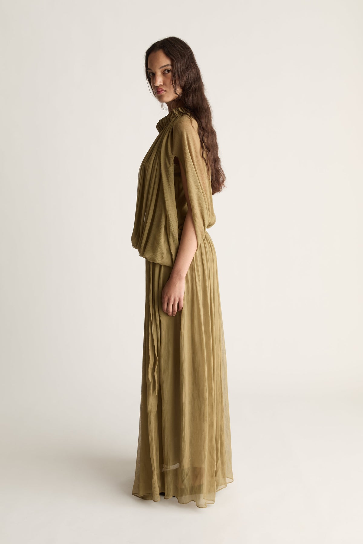 A woman modeling a flowing, draped maxi dress in a muted olive green color, photographed against a plain, light off-white studio background.
