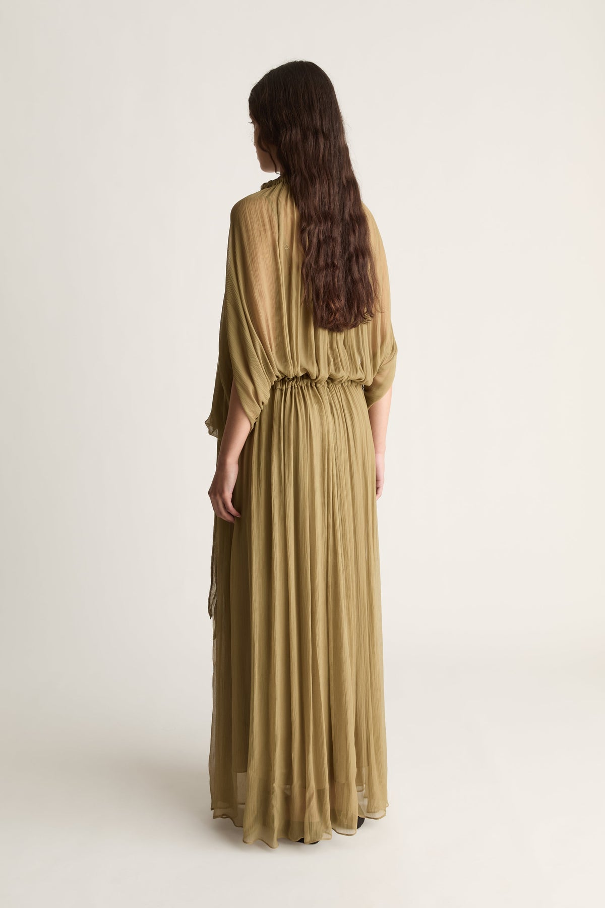 A woman with long dark hair is shown from the back wearing a flowing olive green maxi dress with three-quarter sleeves and a gathered waist, photographed against a plain light beige studio background.