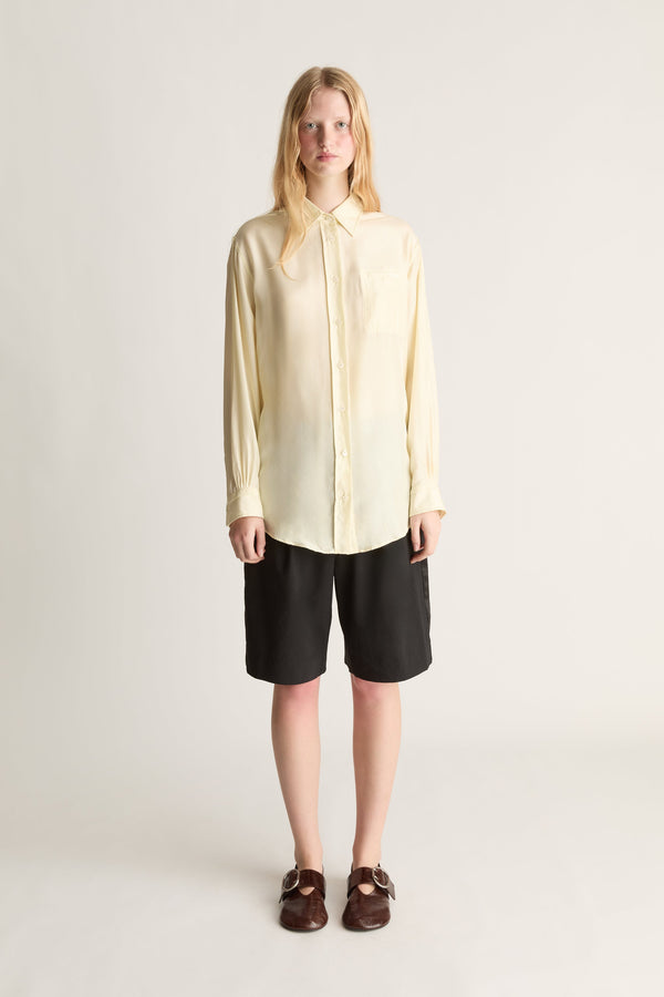 A woman stands against a plain light gray studio background wearing a pale yellow long-sleeve button-up shirt with a tie collar, black shorts, and dark brown loafers.