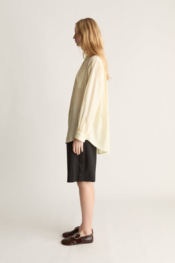A woman shown in profile wearing a loose, oversized cream-colored long-sleeve top, black shorts, and dark brown leather loafers, standing against a plain light gray studio background.