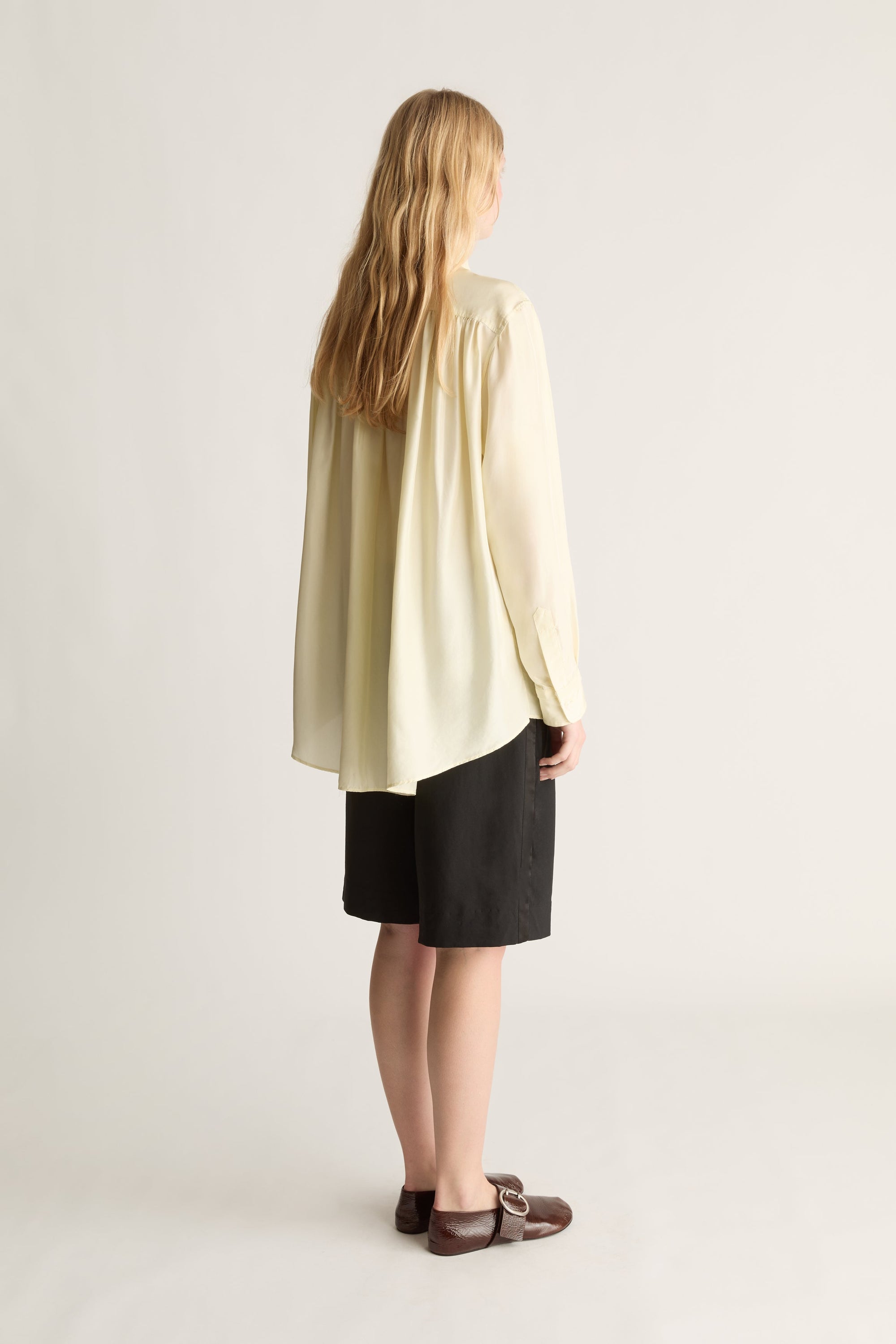 A woman shown from the back wearing a cream-colored long-sleeve blouse, black shorts, and brown flat shoes against a light gray studio background.