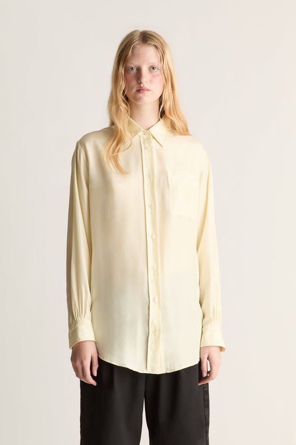 A woman modeling a pale yellow long-sleeve button-up shirt with a collar and chest pocket, paired with black pants, against a plain light gray studio background.