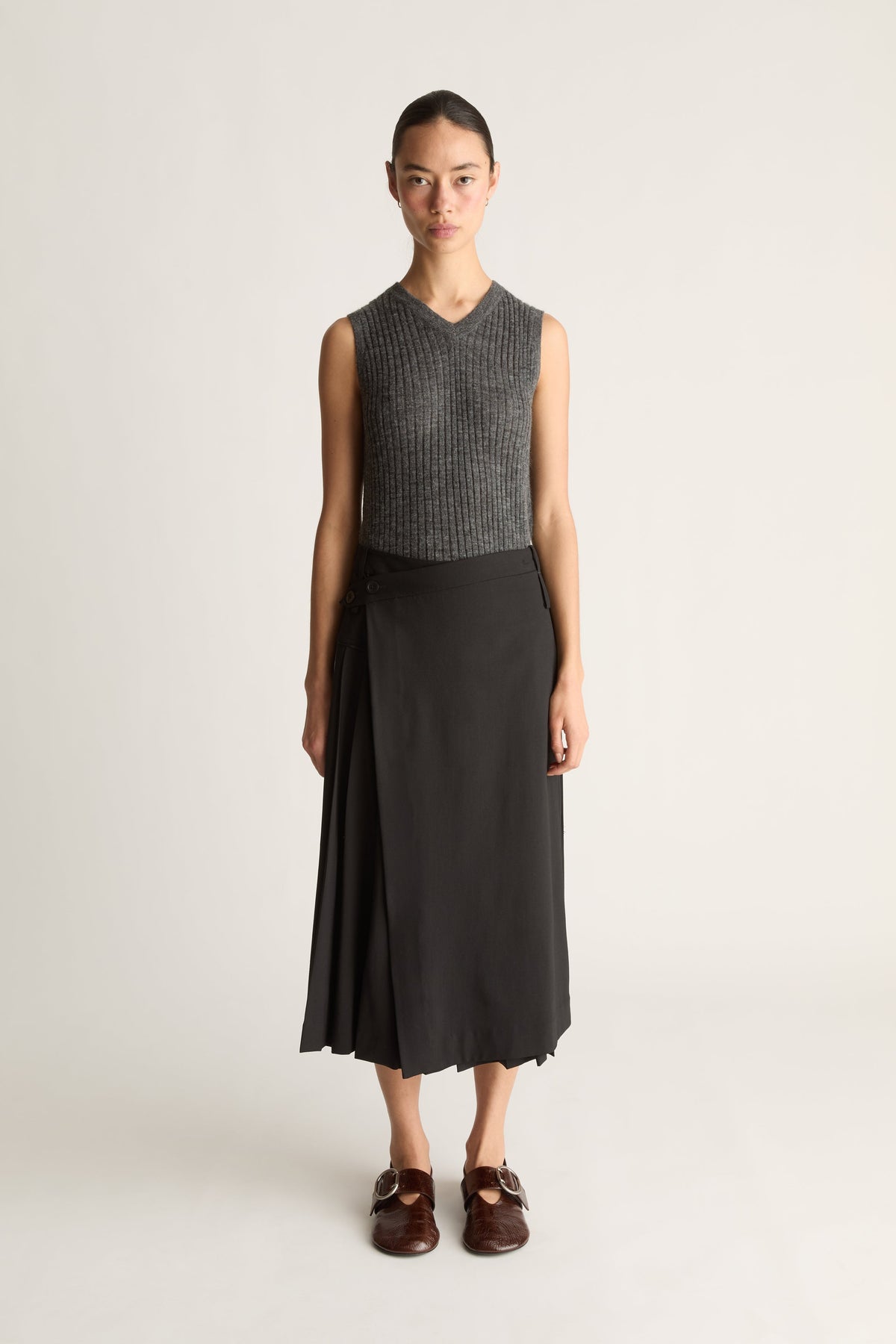 Front view of a woman wearing a knit v-neck grey tank top and a black wool pleated skirt with red shoes against a white studio background.