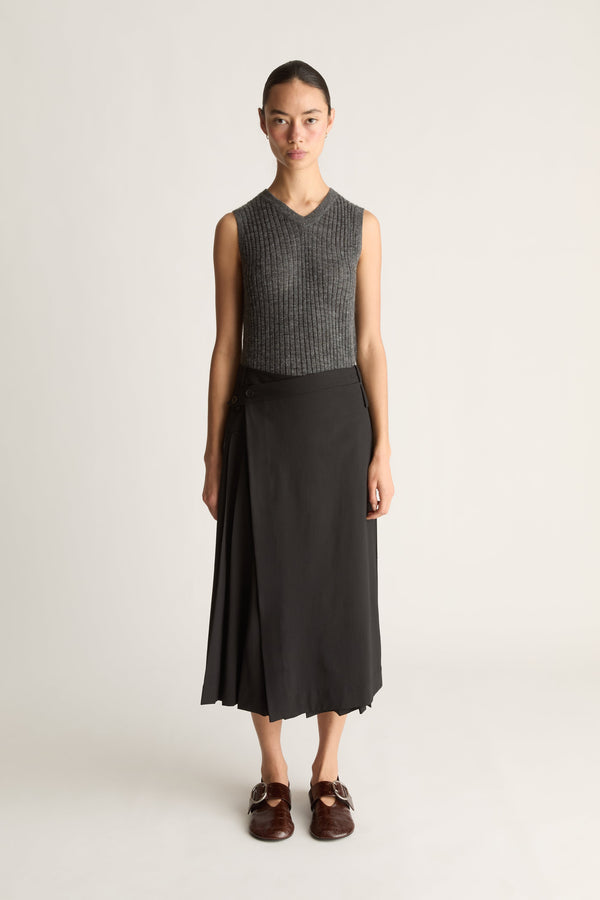 Front view of a woman wearing a knit v-neck grey tank top and a black wool pleated skirt with red shoes against a white studio background.