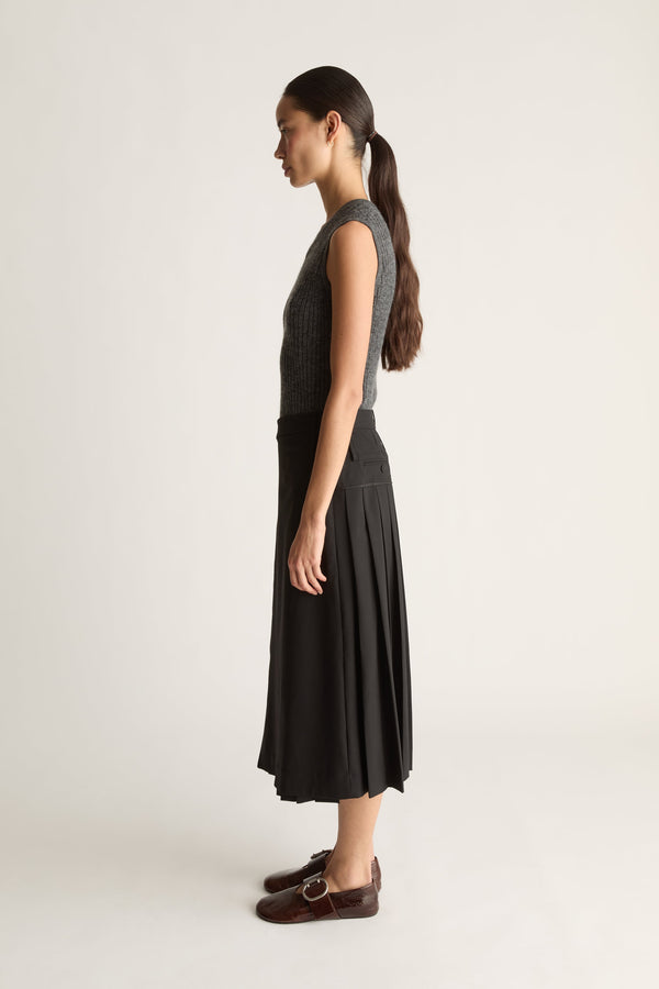 Side profile fo a woman wearing a grey knit tank top and a black pleated wool skirt against a white studio background.