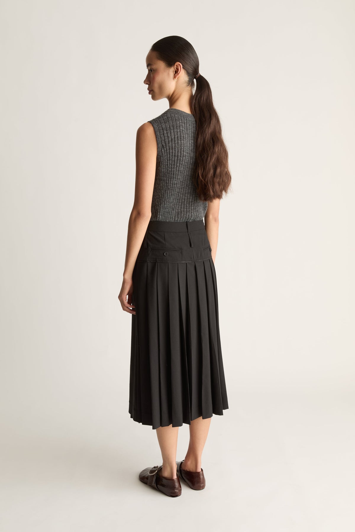 Back view of a woman wearing a grey knit v-neck top and a black wool pleated kilt style skirt agaisnt a white background. 