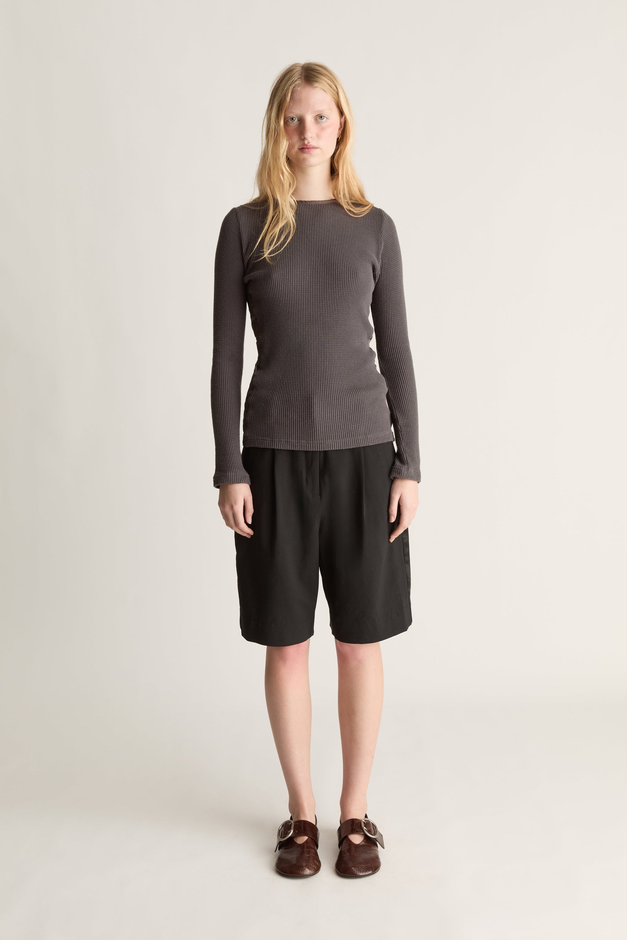 A woman stands against a plain light gray studio background wearing a dark charcoal gray long-sleeve top, black shorts, and brown loafers.