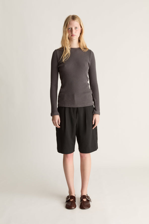 A woman stands against a plain light gray studio background wearing a dark charcoal gray long-sleeve top, black shorts, and brown loafers.