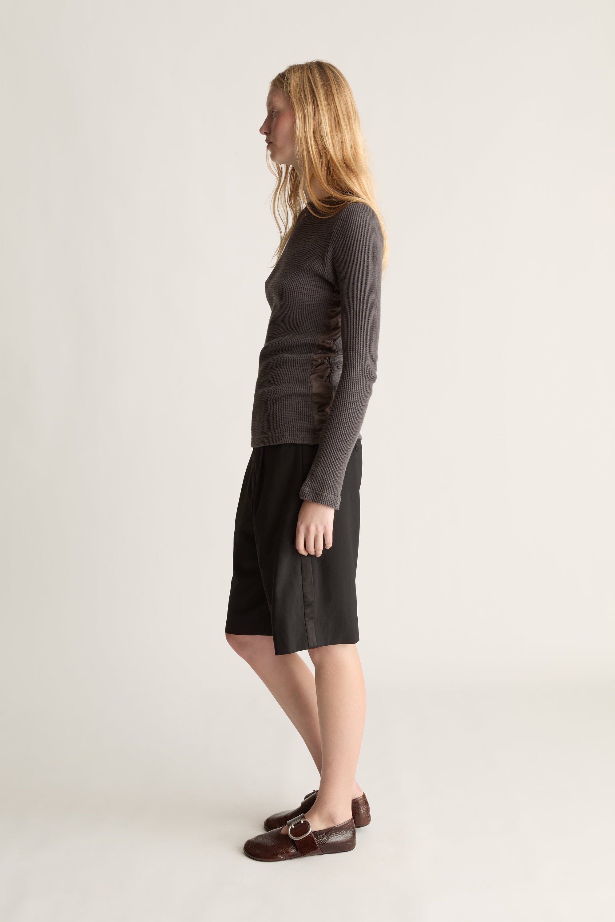 A woman standing in profile wearing a dark charcoal gray long-sleeve top,  black knee-length shorts, and dark brown loafers against a plain light gray studio background.