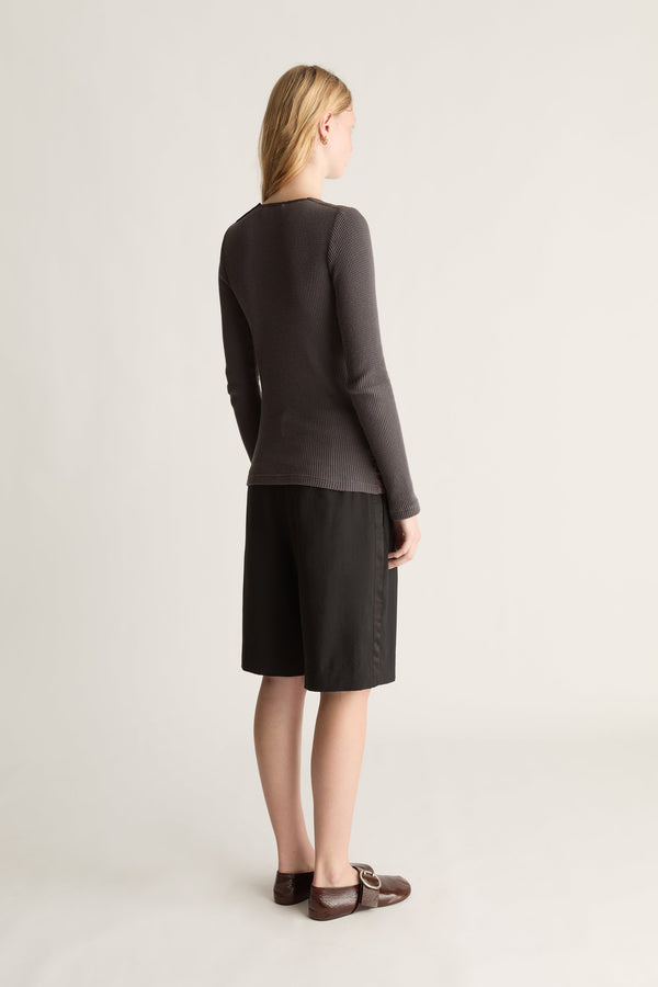 A woman stands with her back to the camera wearing a dark charcoal long-sleeve knit top and black knee-length shorts against a light gray studio background.