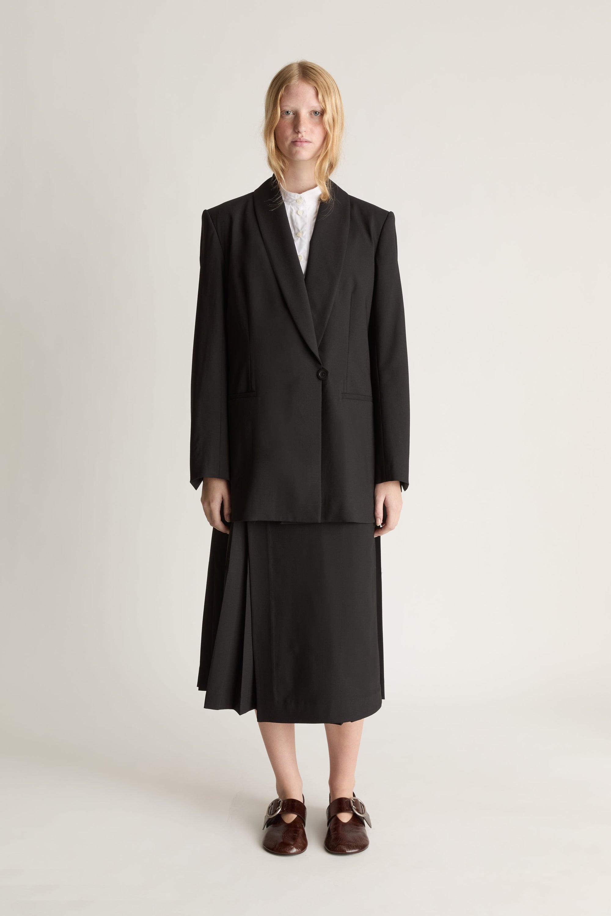 Front view of a woman wearing a black blazer and black pleated midi skirt with red shoes against a white studio background.