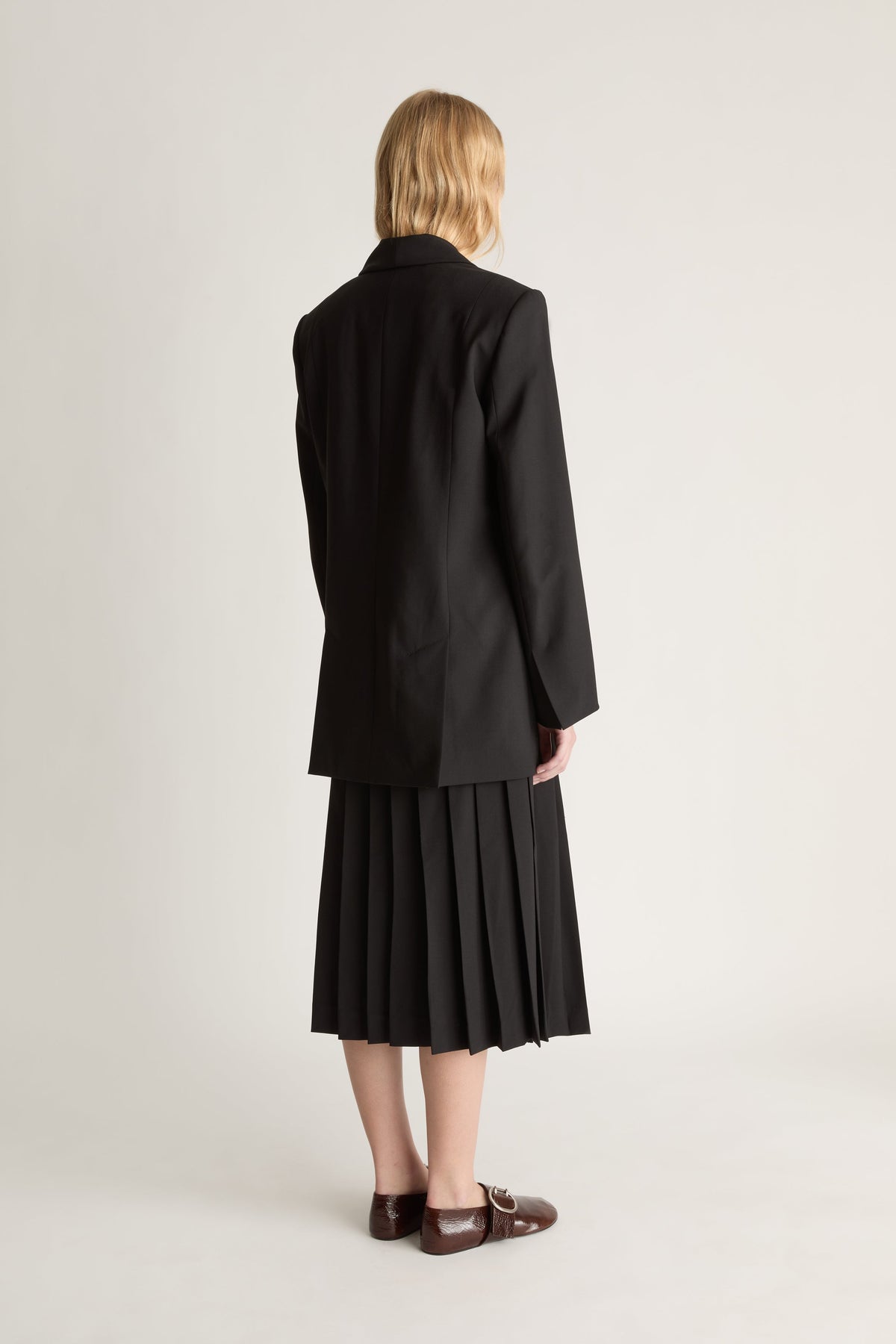 Back view of a person wearing a black blazer and pleated skirt wih red shoes against a white studio background.