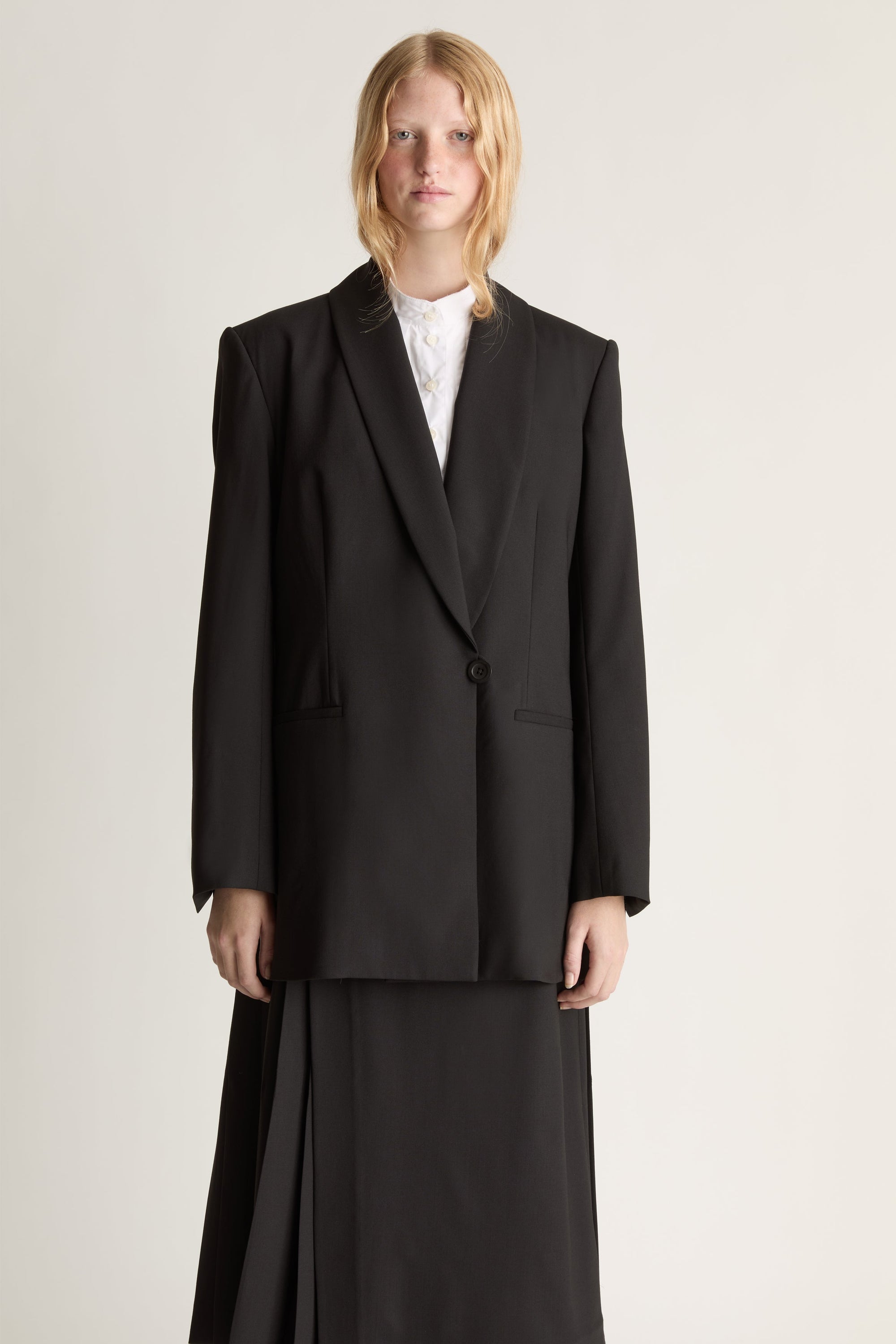 Close up of a woman wearing a black blazer over a white shirt and a black pleated skirt against a studio background.