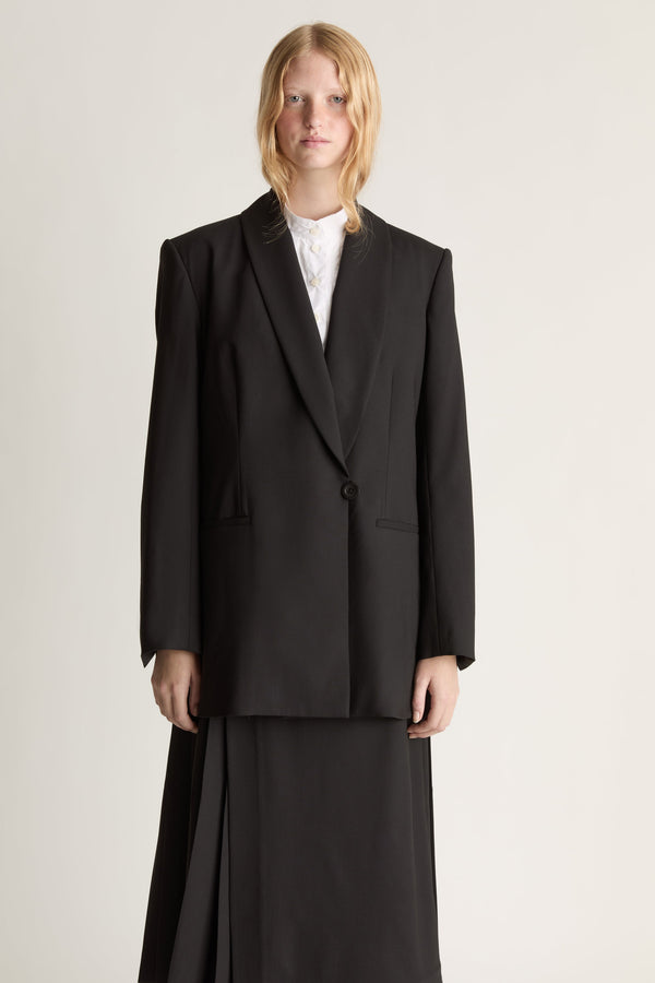 Close up of a woman wearing a black blazer over a white shirt and a black pleated skirt against a studio background.