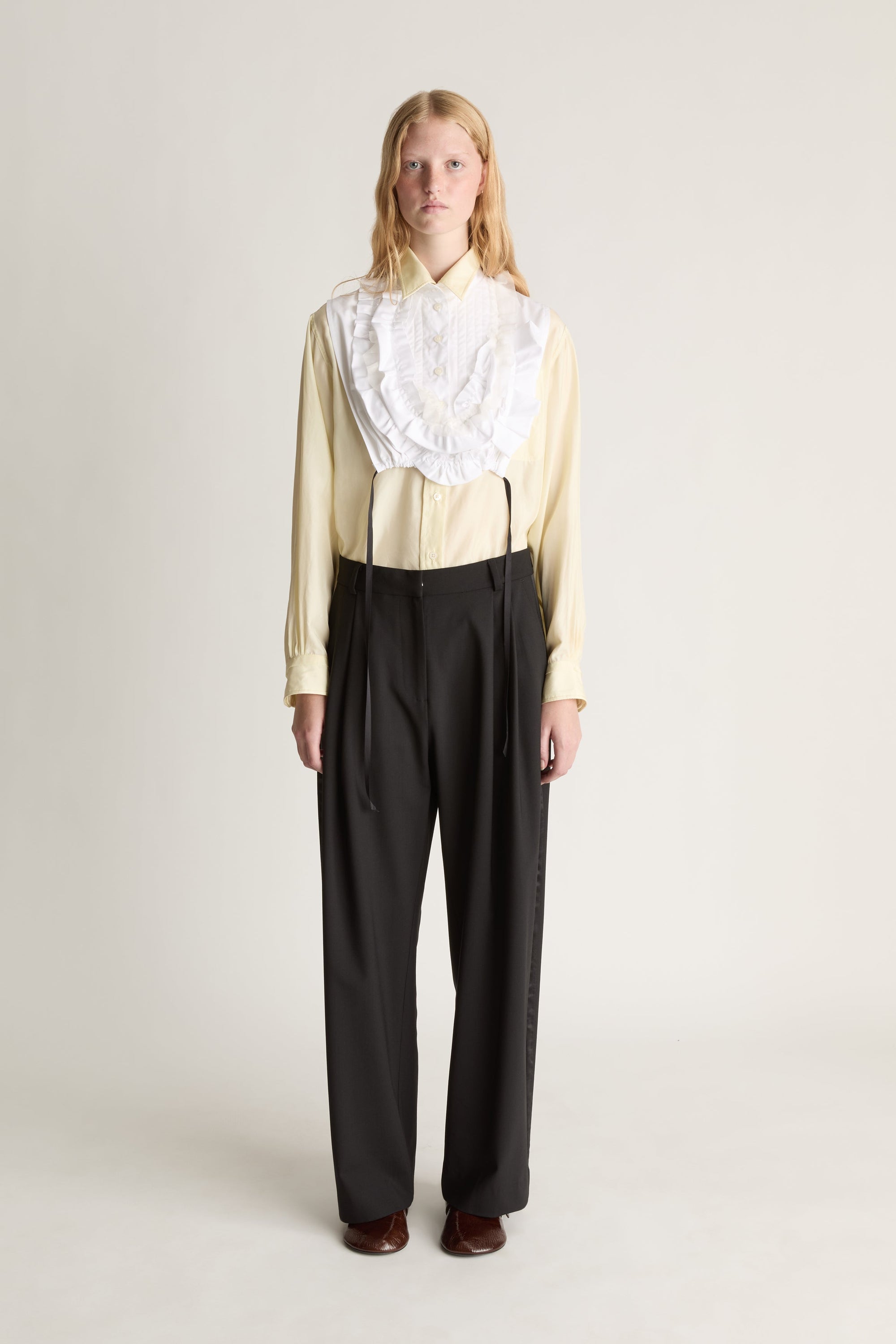 Person wearing a white frill dickey over a yellow blouse and black pants on a plain background