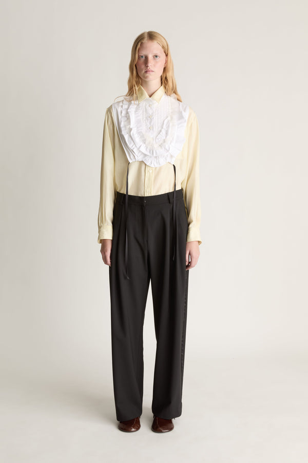 Person wearing a white frill dickey over a yellow blouse and black pants on a plain background