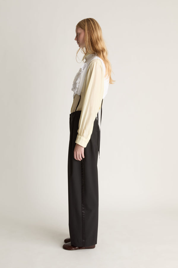 Model standing in profile wearing a yellow blouse with a white frill vest over the top and black trousers against a white studio background.