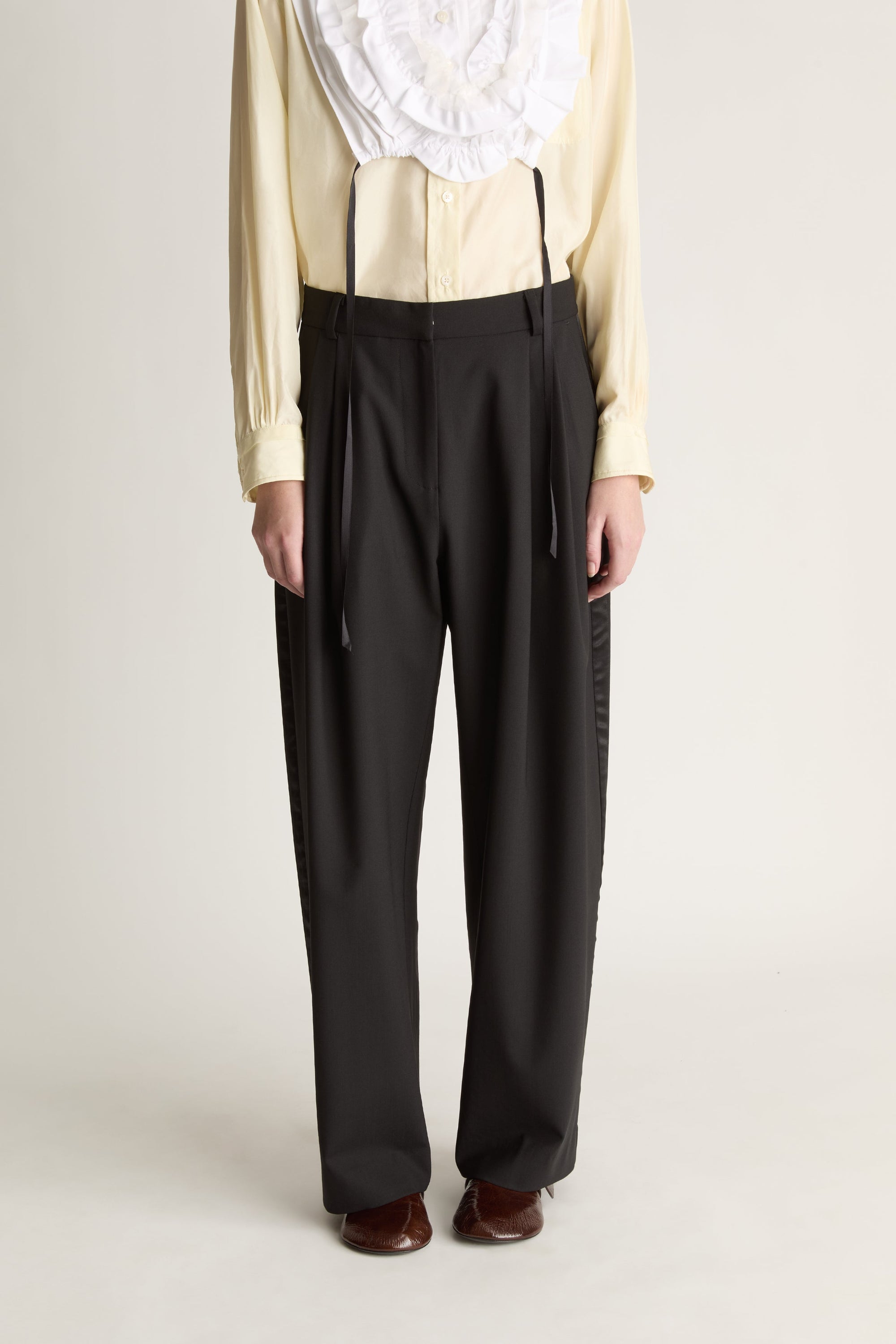 Close up image of a person wearing a white frill dickey over a yellow blouse and black pants on a plain background