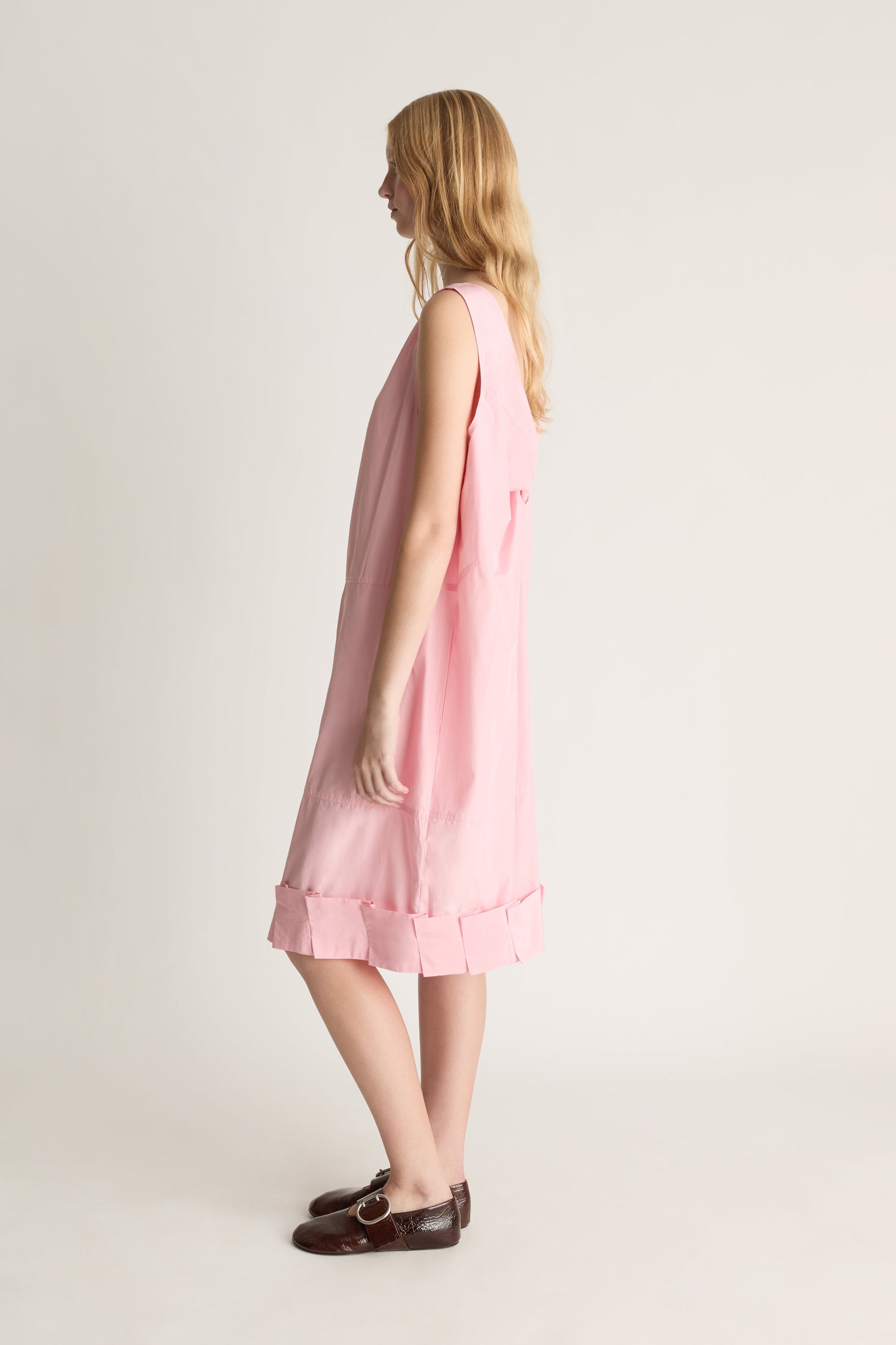Woman standing in profile wearing a silk pink midi dress with a pleated detail along the hemline, against a white studio background. 