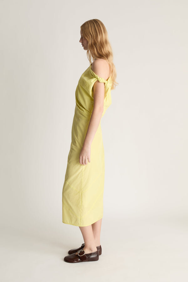 Side view of a woman wearing an off the shoulder yellow silk dress with red shoes against a white studio background. 