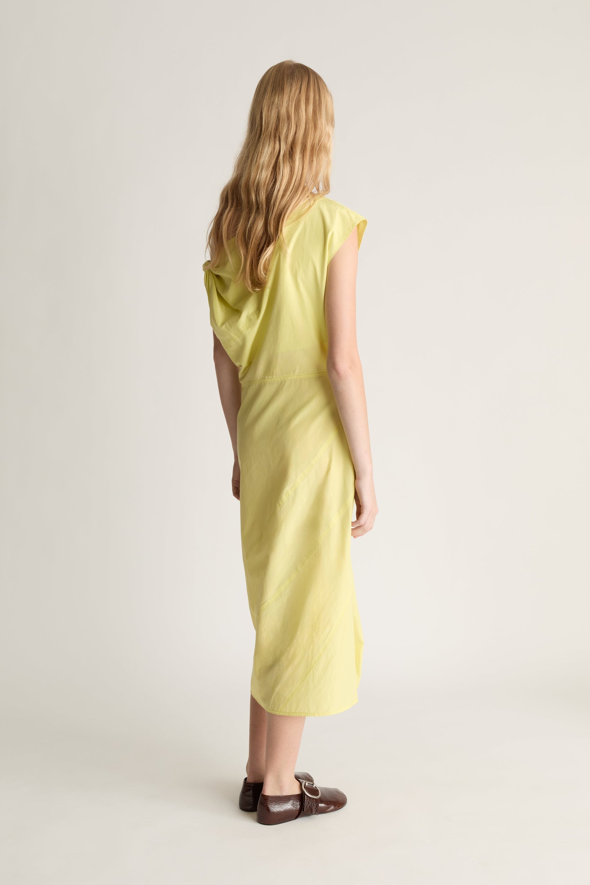 Back view of a woman wearing a light yellow midi dress and dark brown loafers, standing against a plain light gray studio background.