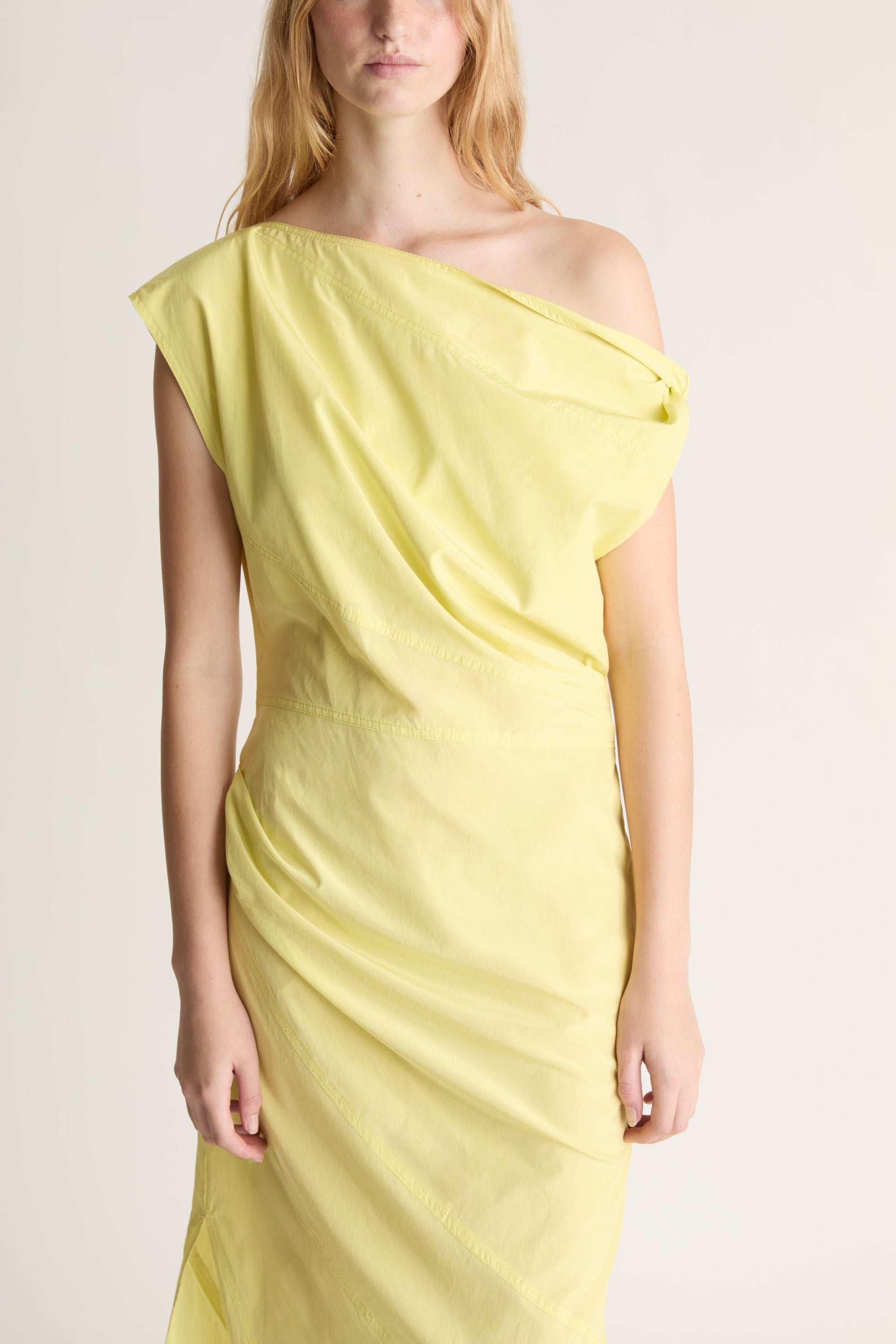 Close up of a woman wearing a yellow one-shoulder dress against a plain background