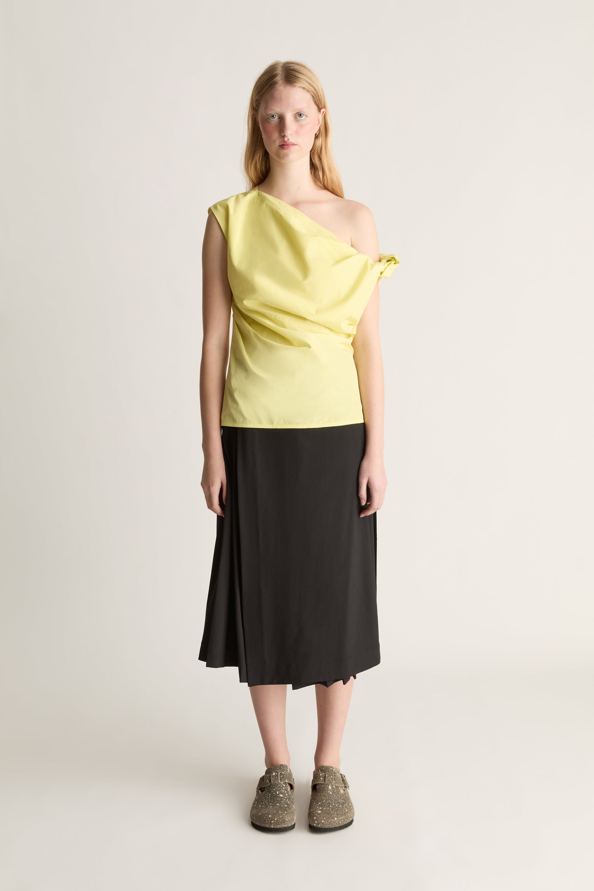A model stands against a light beige studio background wearing a pale yellow asymmetrical one-shoulder top with a tie detail at the shoulder, paired with a black midi skirt and brown lace-up shoes.