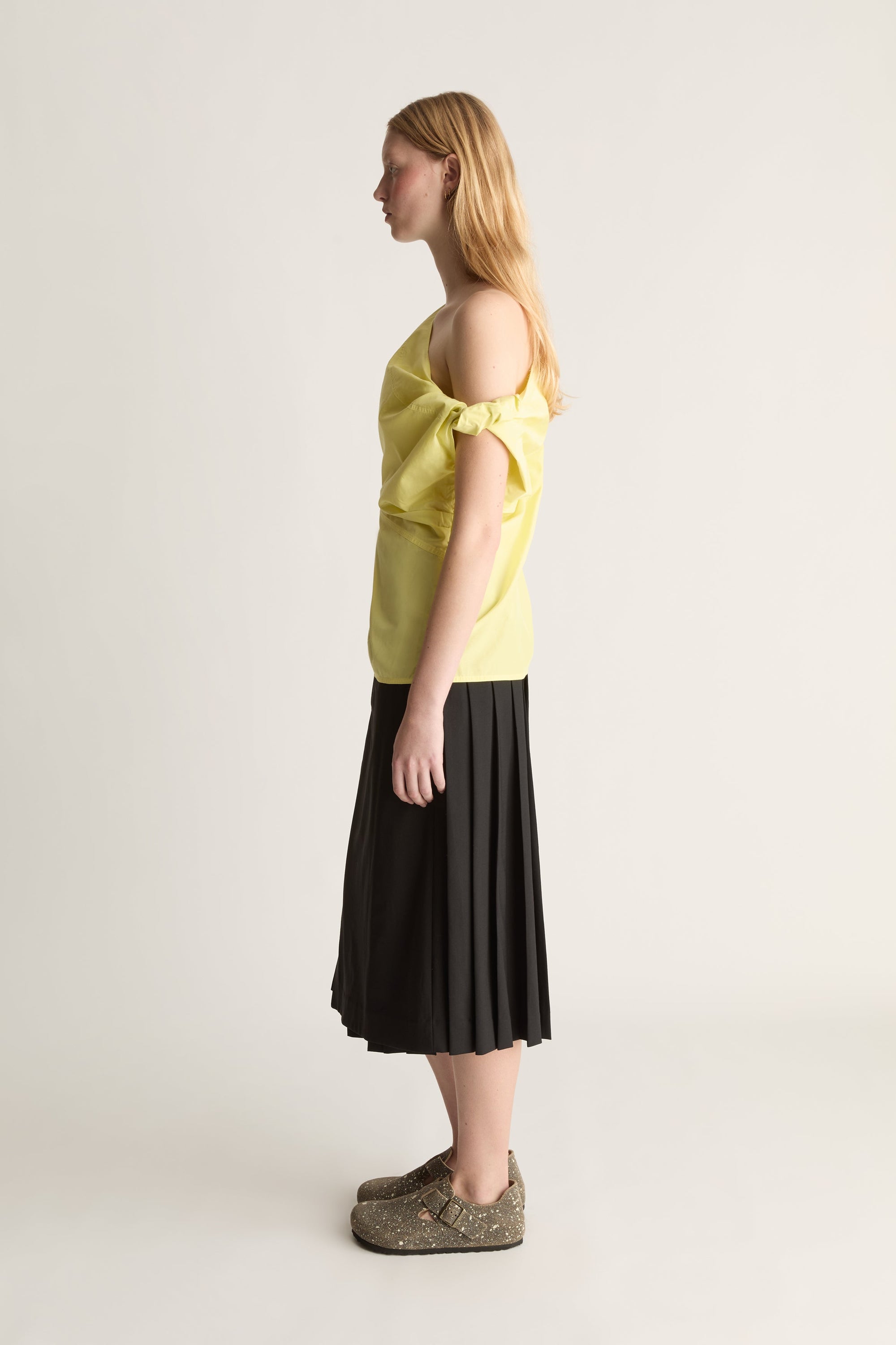 A woman stands in profile wearing a bright yellow cold-shoulder top with ruffled sleeves, a black pleated midi skirt, and textured brown slip-on shoes against a plain light gray studio background.