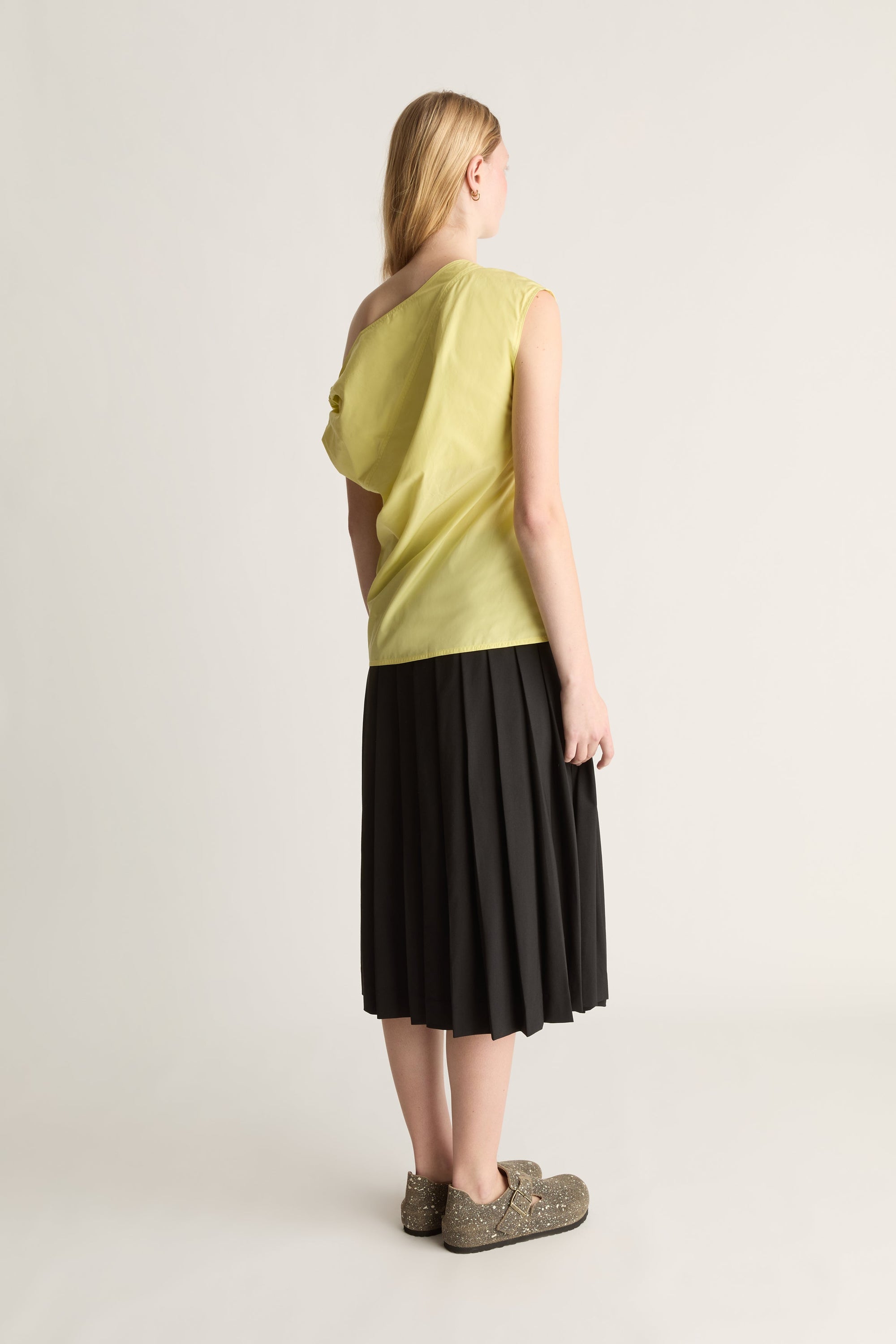 A woman stands with her back to the camera wearing a sleeveless lime green top and a black pleated midi skirt, paired with brown textured slip-on shoes, against a plain light gray studio background.