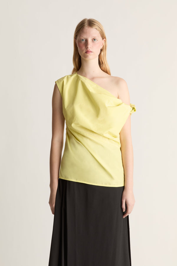 Close-up of a model wearing a pale yellow asymmetrical one-shoulder top with a black skirt, standing against a light gray studio background.