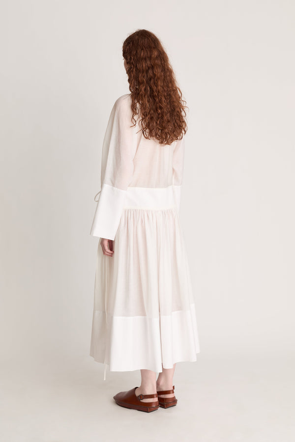 A woman wearing a long white dress with a tie detail, on a plain background