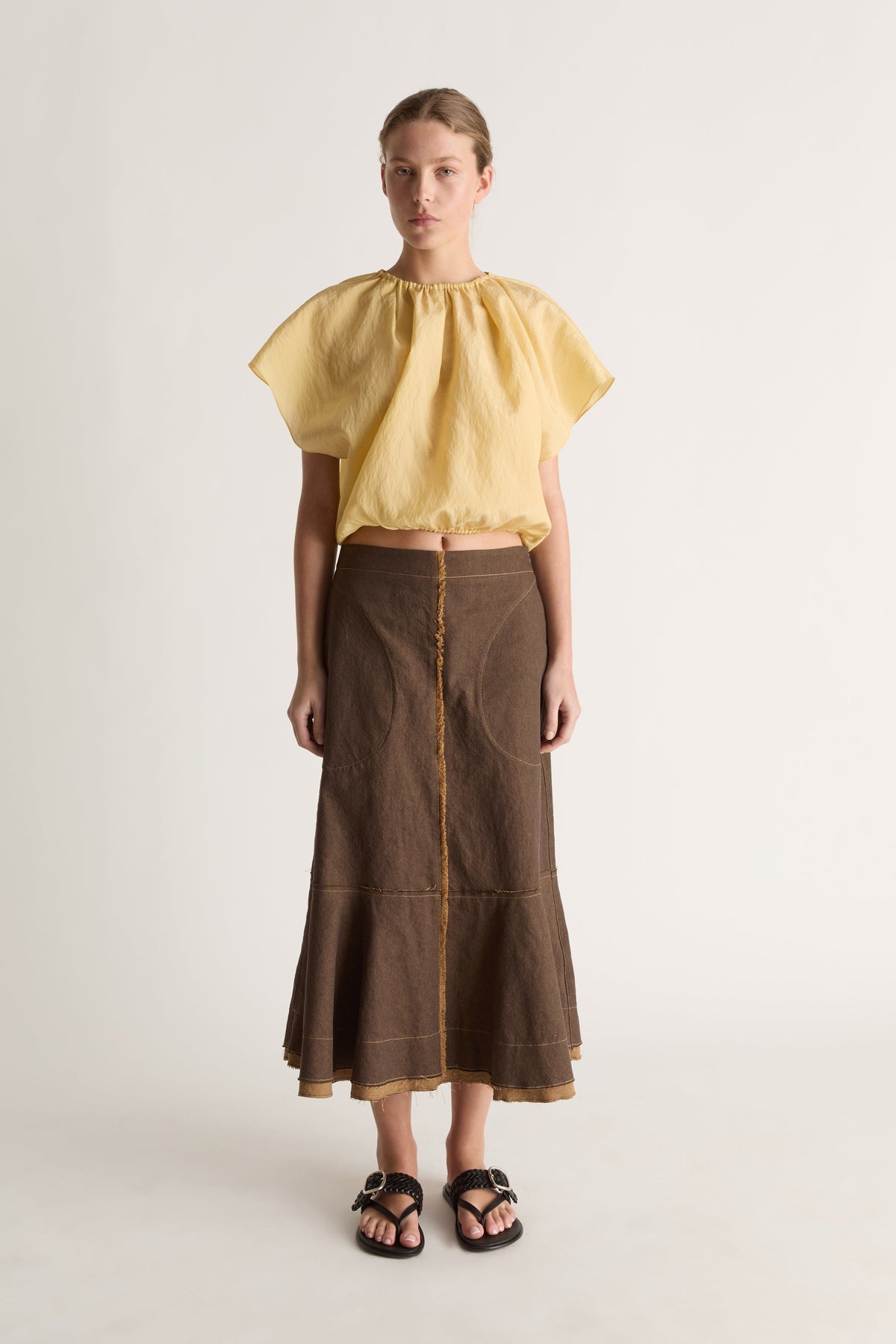 Front view of a woman wearing a yellow gathered cropped top and brown flared midi skirt against a white studio background