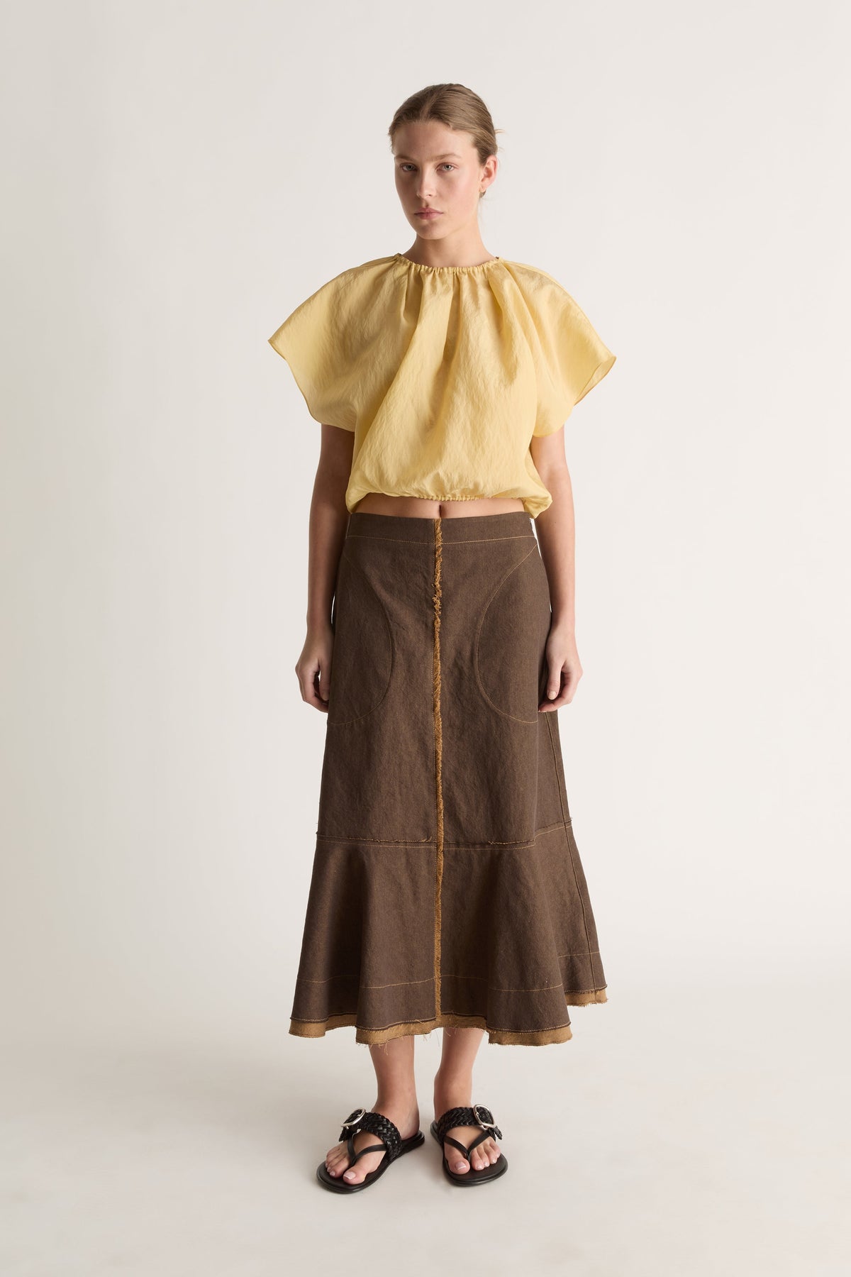 Straight-on view of a woman with hair pulled back in a short-sleeve yellow blouse, brown fluted-hem skirt and black slides