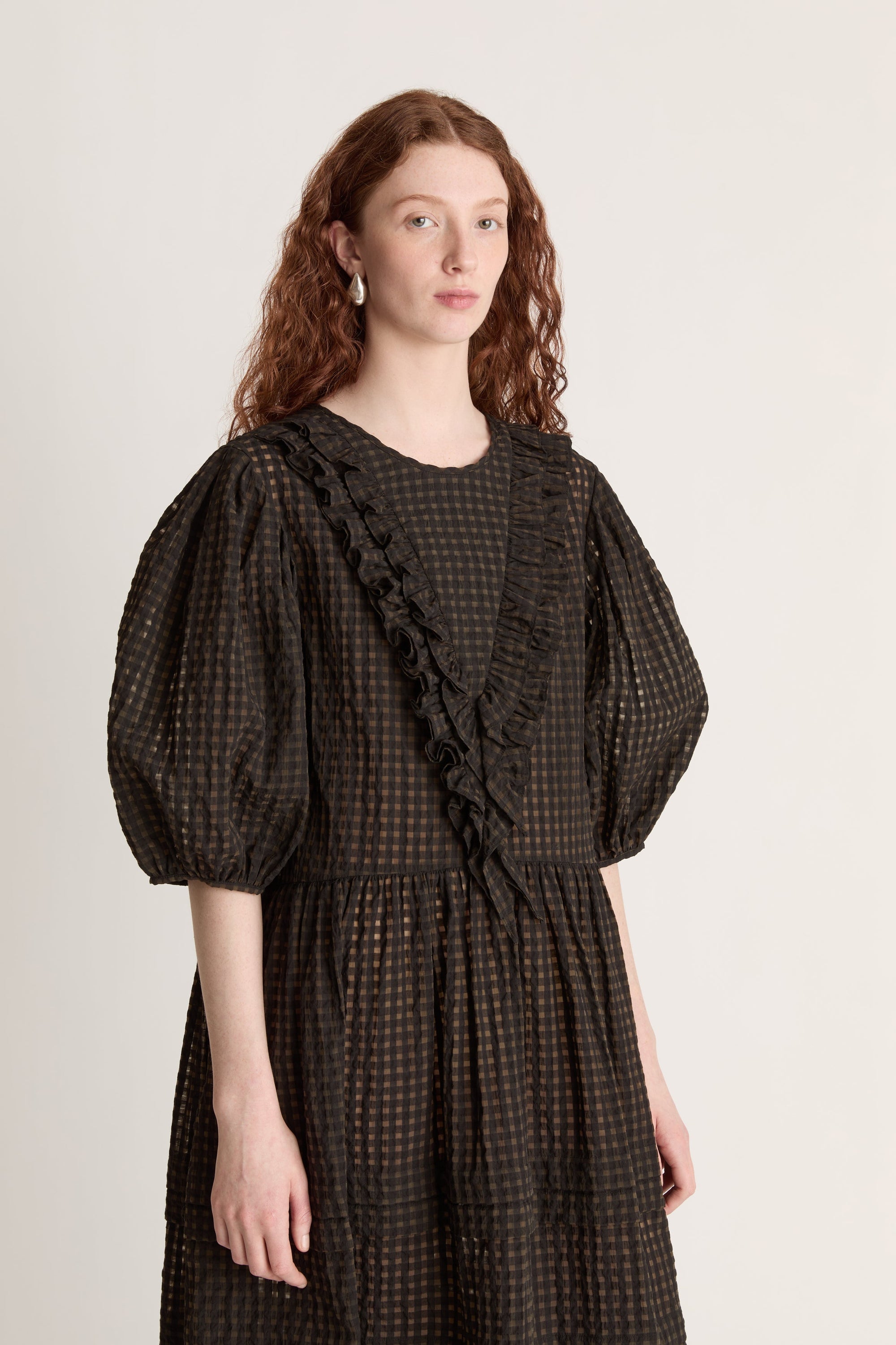 Woman wearing a dark checkered dress  with puffy sleeves with ruffled details on a white background