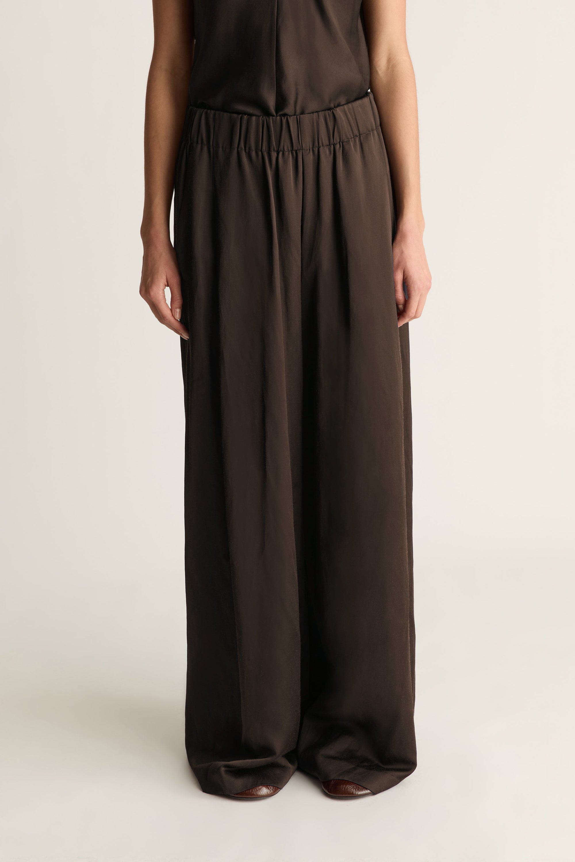 A model wearing dark brown wide-leg trousers with an elastic waistband, shown from the chest down against a plain light background.