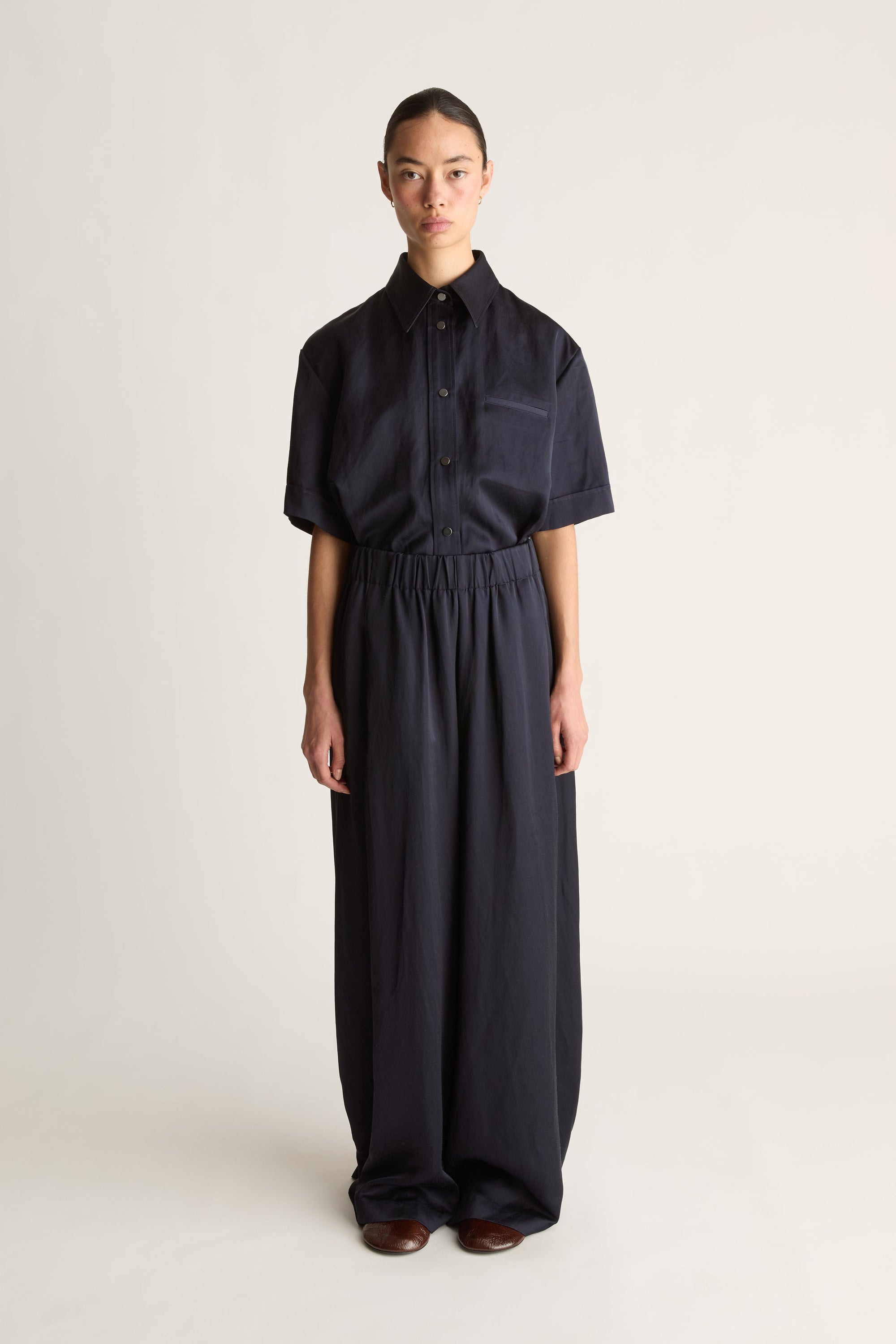 Front view of a woman wearing a navy short-sleeved shirt and elasticated wide-leg trousers, shot against a white studio background.