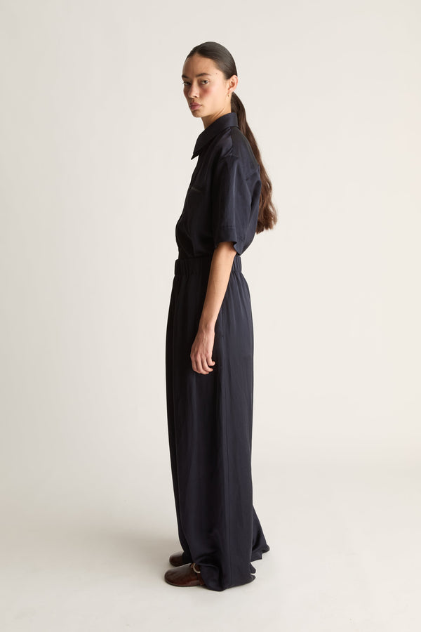 A model stands in profile wearing a dark navy blue long-sleeve shirt and matching wide-leg trousers, photographed against a plain light beige studio background.