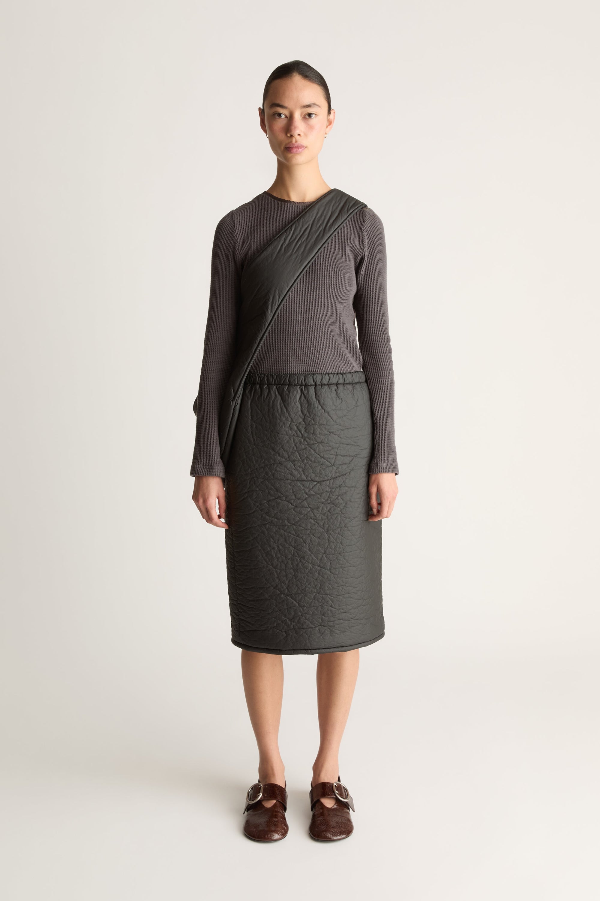 A model stands against a light gray studio background wearing a dark charcoal gray long-sleeve shirt and a black puff material skirt with a matching black cross body bag/
