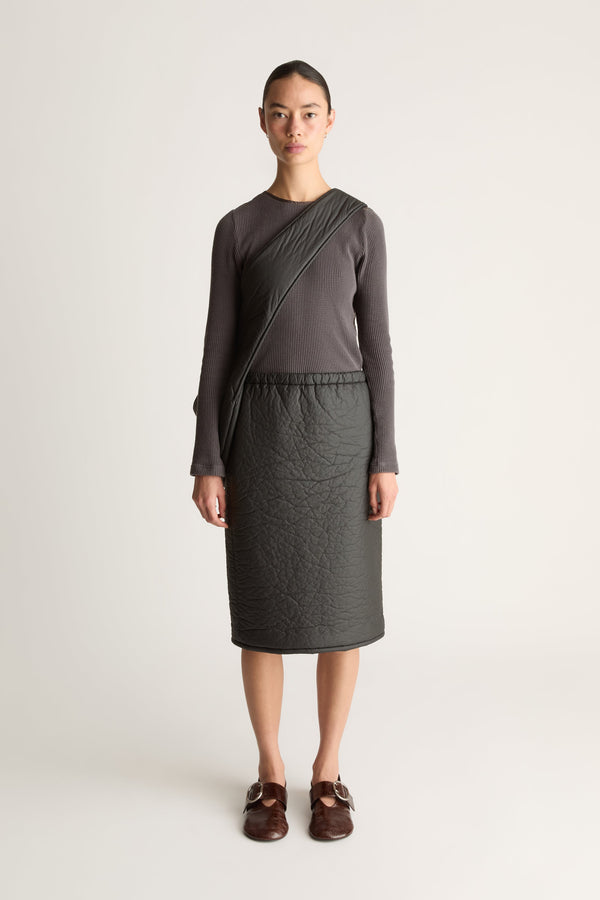 A model stands against a light gray studio background wearing a dark charcoal gray long-sleeve shirt and a black puff material skirt with a matching black cross body bag/
