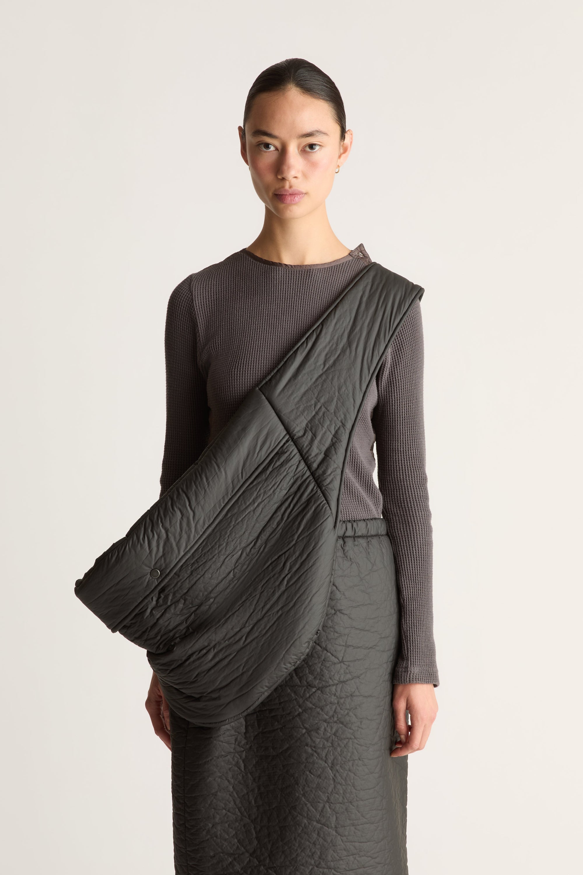 A model wearing a dark gray ribbed long-sleeve top and black skirt, with a large, draped dark gray quilted fabric bag or wrap styled over her shoulder, set against a plain light gray studio background.