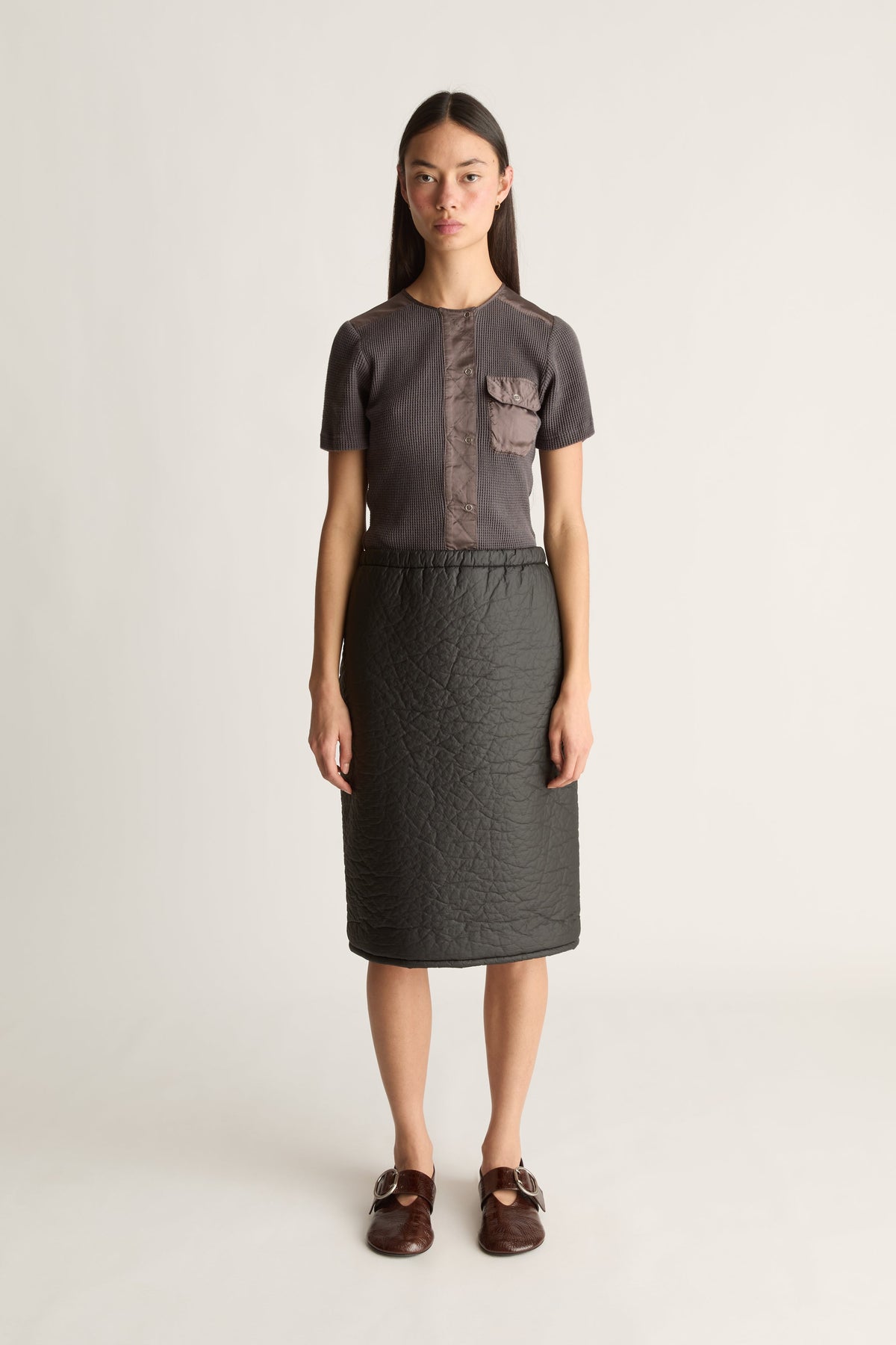 A model stands against a light gray studio background wearing a dark gray short-sleeve top with a textured vertical chest panel and matching dark gray skirt, paired with brown loafers.